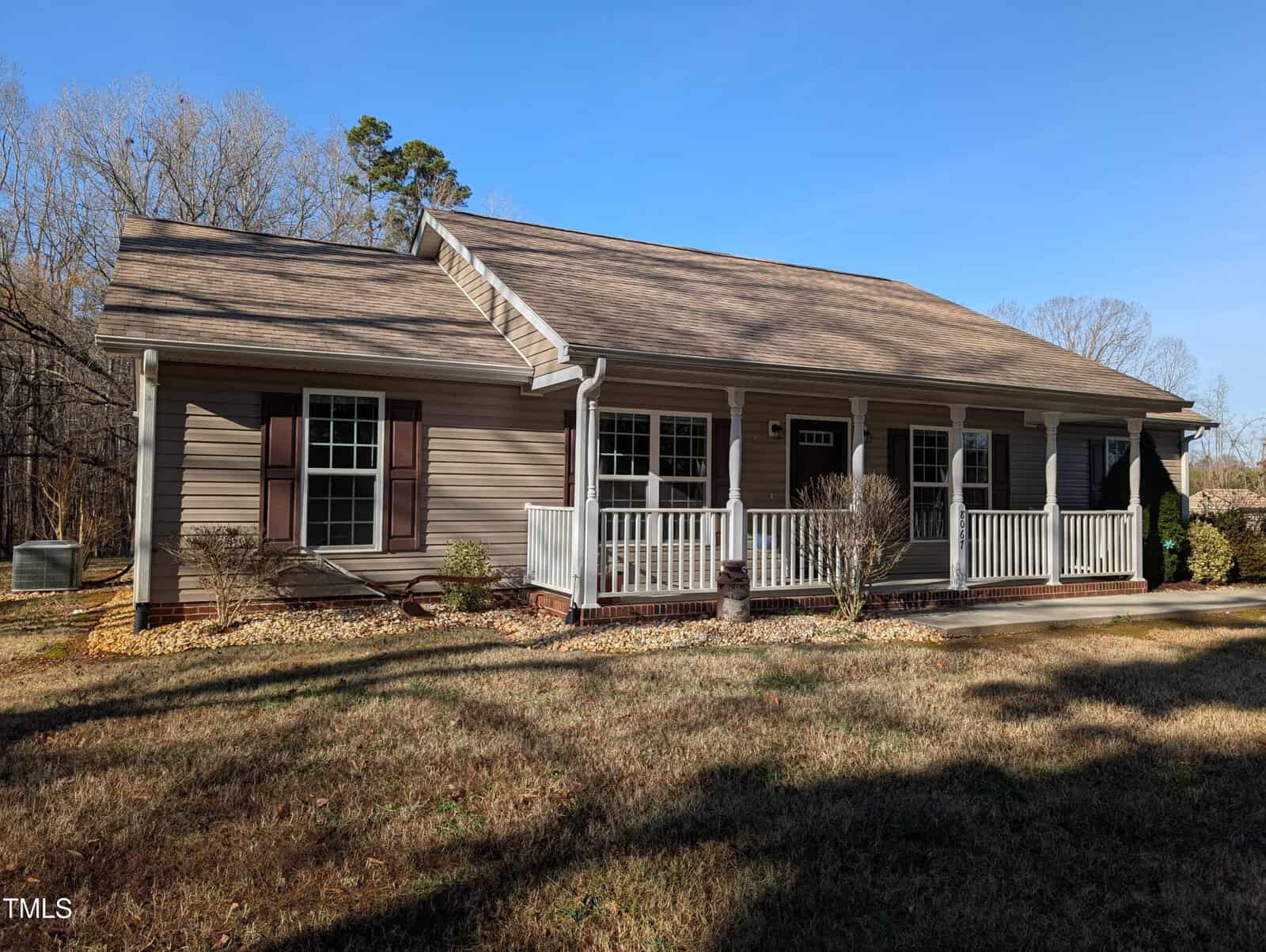 This charming single-story house on 8067 NC Hwy 96 features a gabled roof and a covered front porch. Its tan exterior is accented with white trim and dark shutters. The grassy yard, dotted with shrubs and trees, completes this peaceful Oxford setting under a clear blue sky.