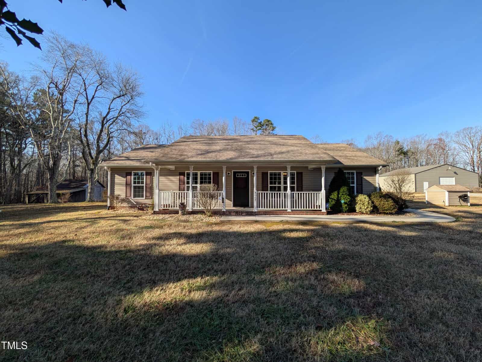 A single-story house with a beige exterior and a gray roof sits on a grassy lawn. It has a front porch with white railings. There are trees in the background and a clear blue sky above. A smaller structure is visible to the right.