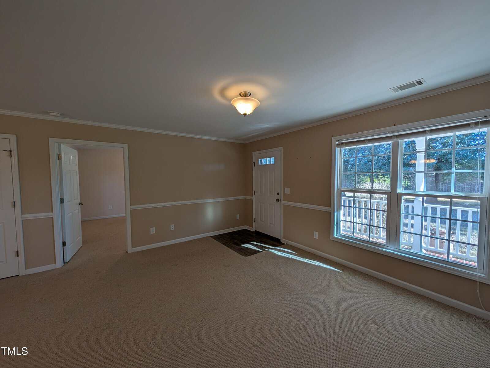 A spacious, empty room with beige walls and carpeted flooring. There is a ceiling light fixture, a large window letting in natural light, and an open doorway leading to another room. A closed door and a small entryway with a mat are also visible.