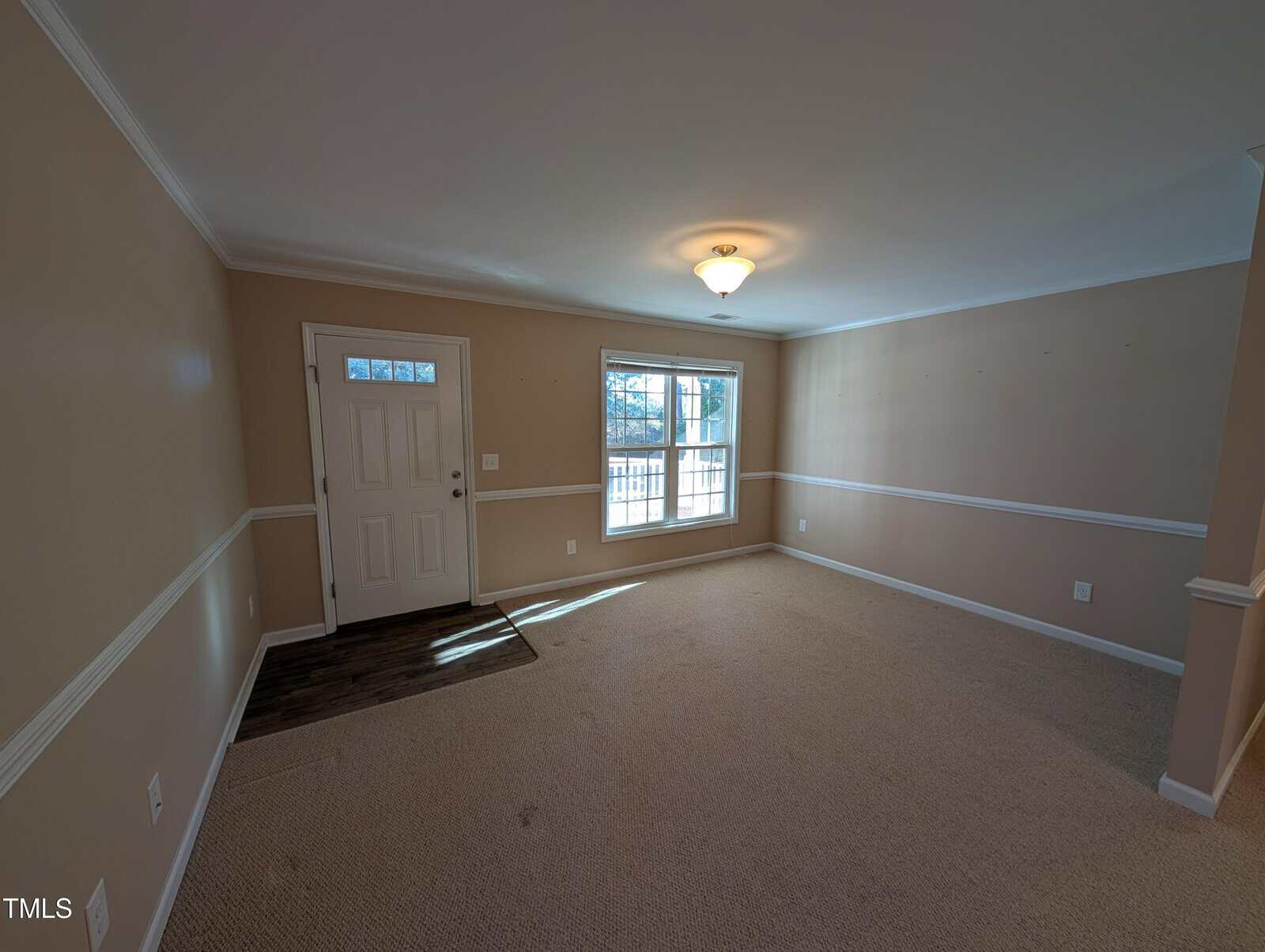 An empty room with beige walls and carpeted floor. A window with a view of trees lets in natural light. A single ceiling light fixture is centered. There is a white door on the left with a small mat in front of it. Wainscoting decorates the walls.