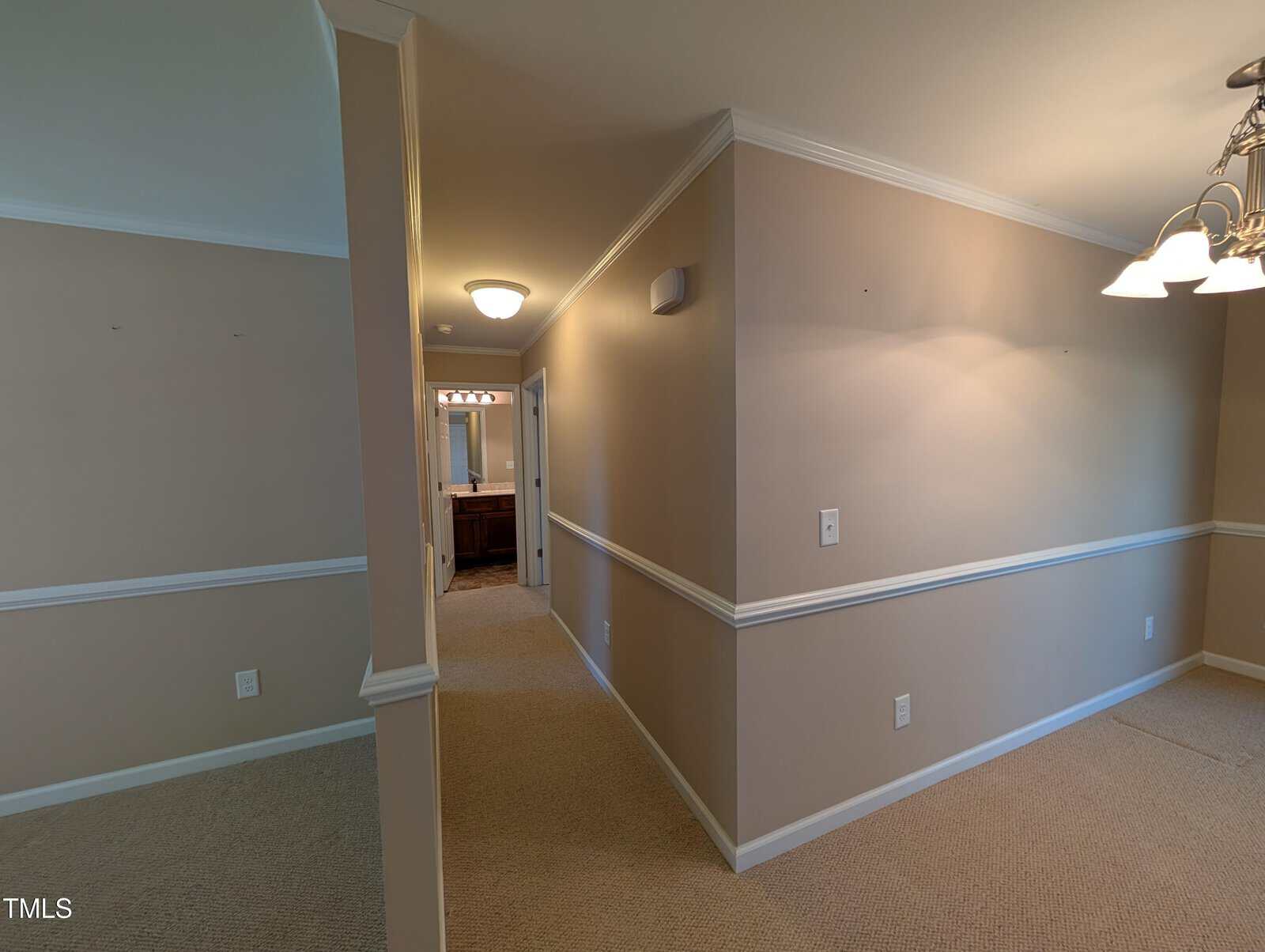 Interior view of a hallway leading to a bathroom. The walls are painted beige with white trim and wainscoting. The ceiling is fitted with a light fixture, and the floor is carpeted. A chandelier is visible on the right side.