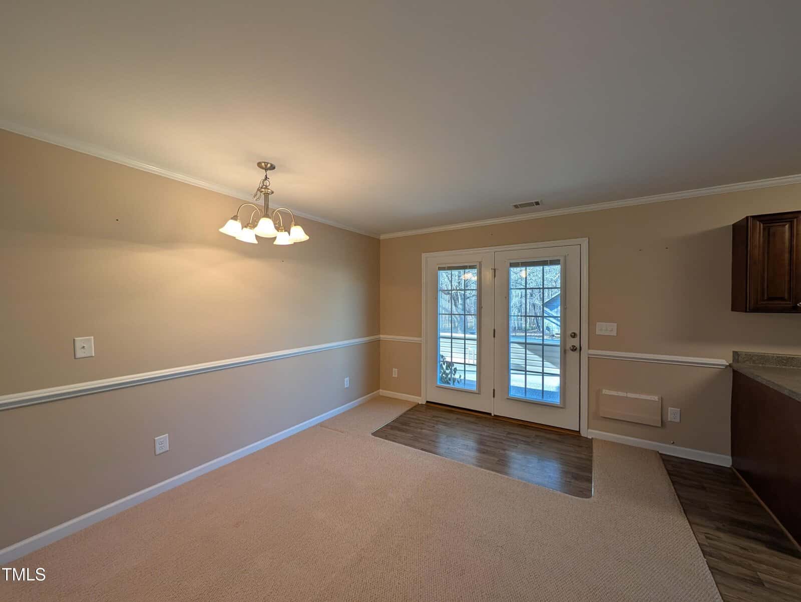 A room with beige walls and carpet features a ceiling light fixture and double glass doors leading outside. Wooden flooring is near the entrance, and a small section of a kitchen countertop is visible on the right.