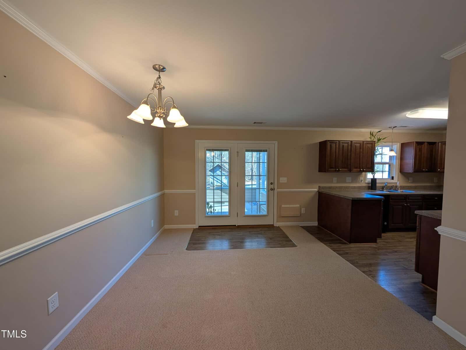 A spacious, empty room with beige walls, a carpeted area, and a hanging light fixture. Double doors with glass panels lead outside. The room opens into a kitchen area with wooden cabinets and a window.