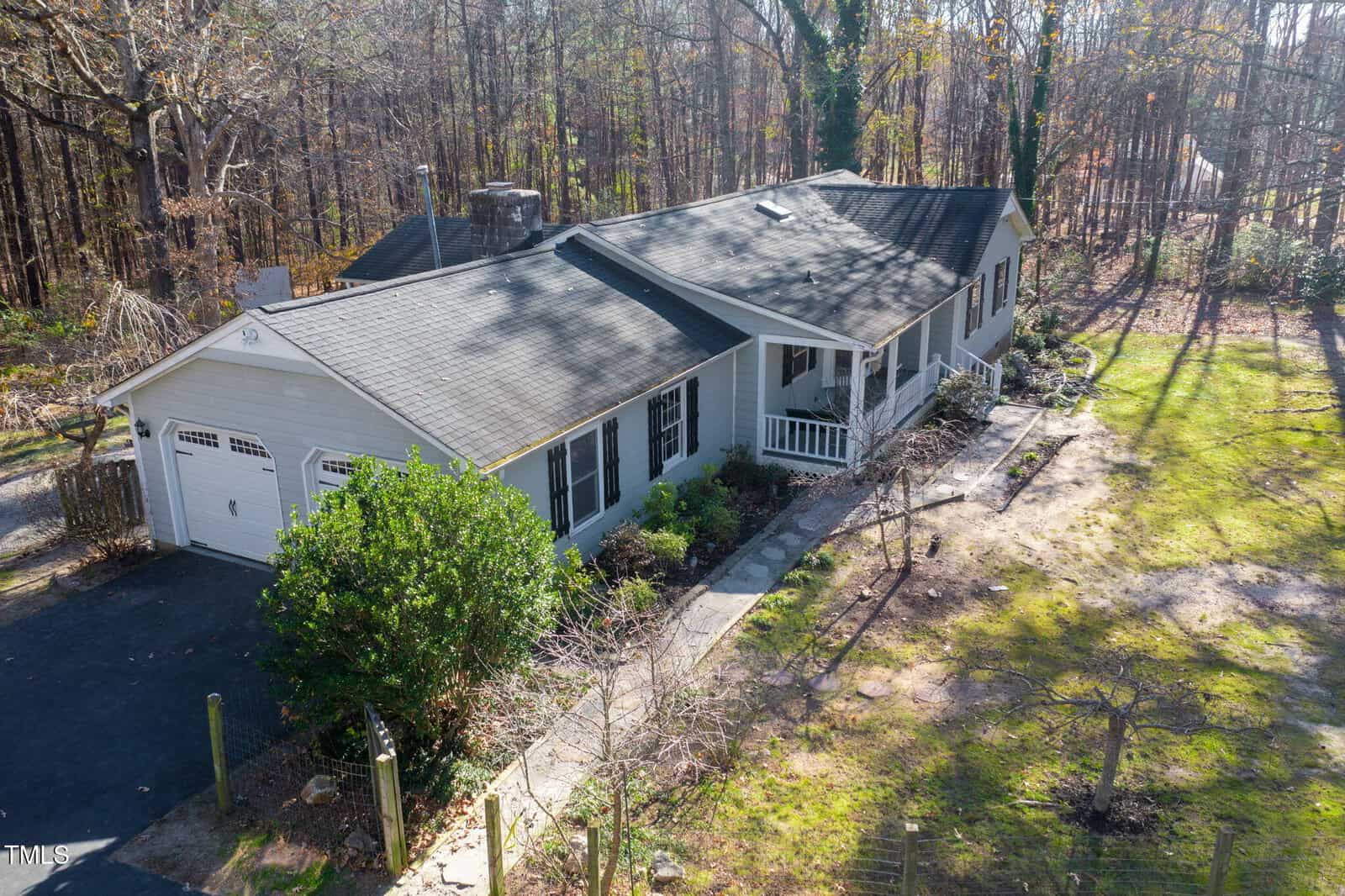A single-story gray house with a gabled roof, attached two-car garage, covered front porch, and surrounding trees on a wooded lot, viewed from above during daylight.