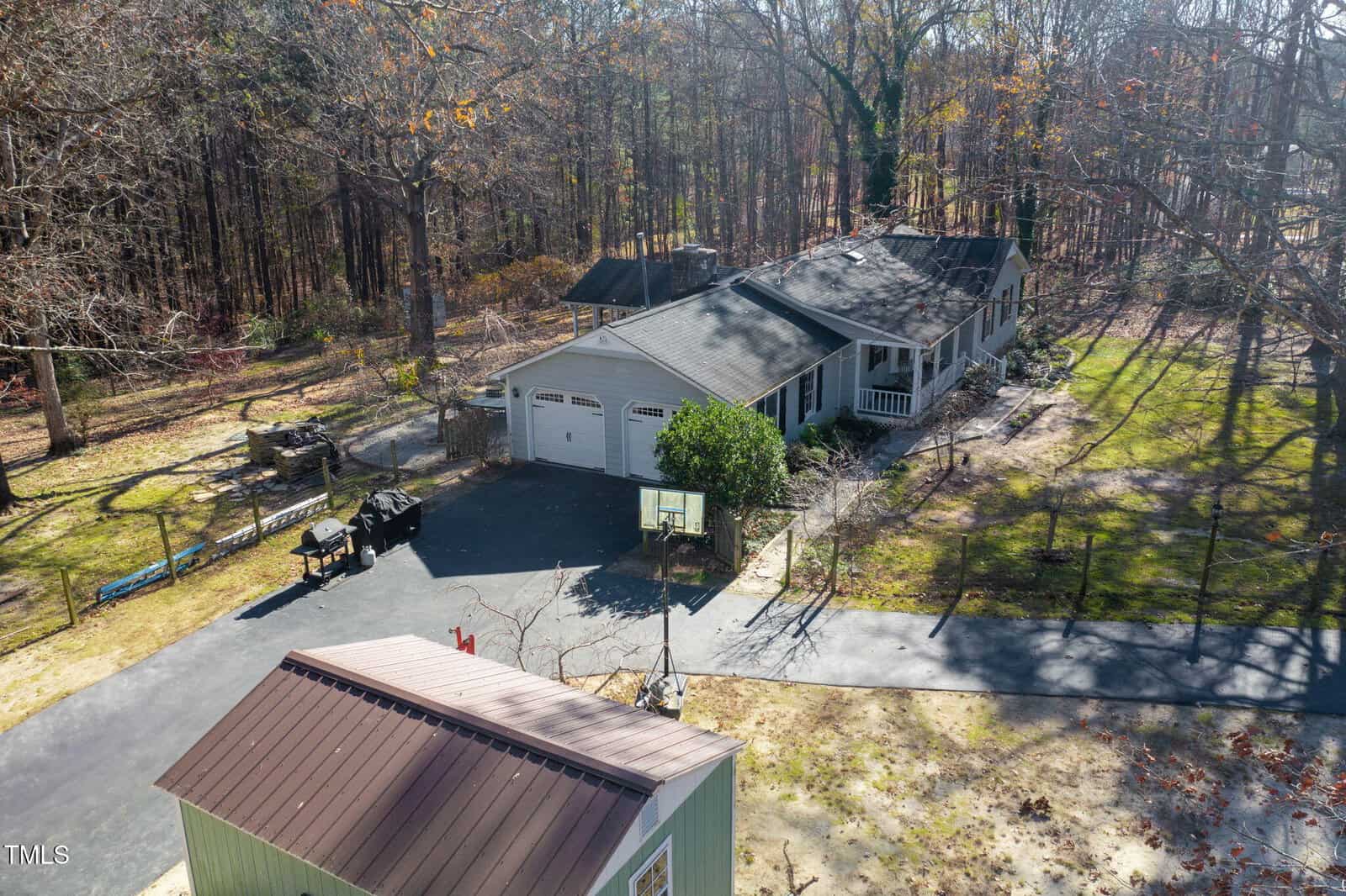 A single-story house with an attached two-car garage, front porch, and paved driveway sits among tall trees. A small outbuilding and a utility trailer are visible in the yard. The area has scattered leaves and light shade.