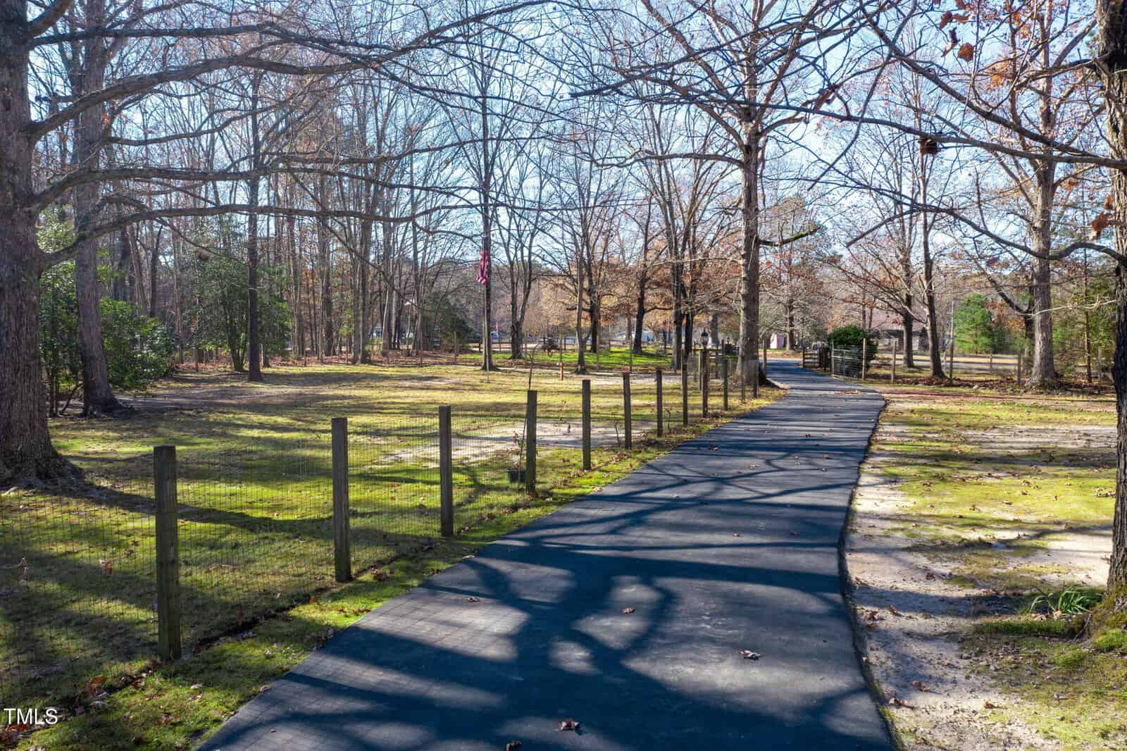 A paved walking path runs through a park with bare trees, a wooden fence on the left side, and patches of grass and sunlight on the ground. The sky is clear and blue, with some scattered leaves remaining on the trees.