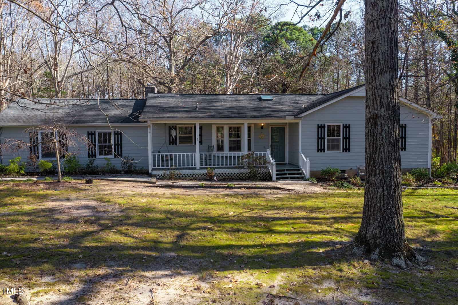 A single-story gray house with white trim and black shutters, featuring a covered front porch with railings. The yard has patchy grass, some trees, and is surrounded by woods in the background.