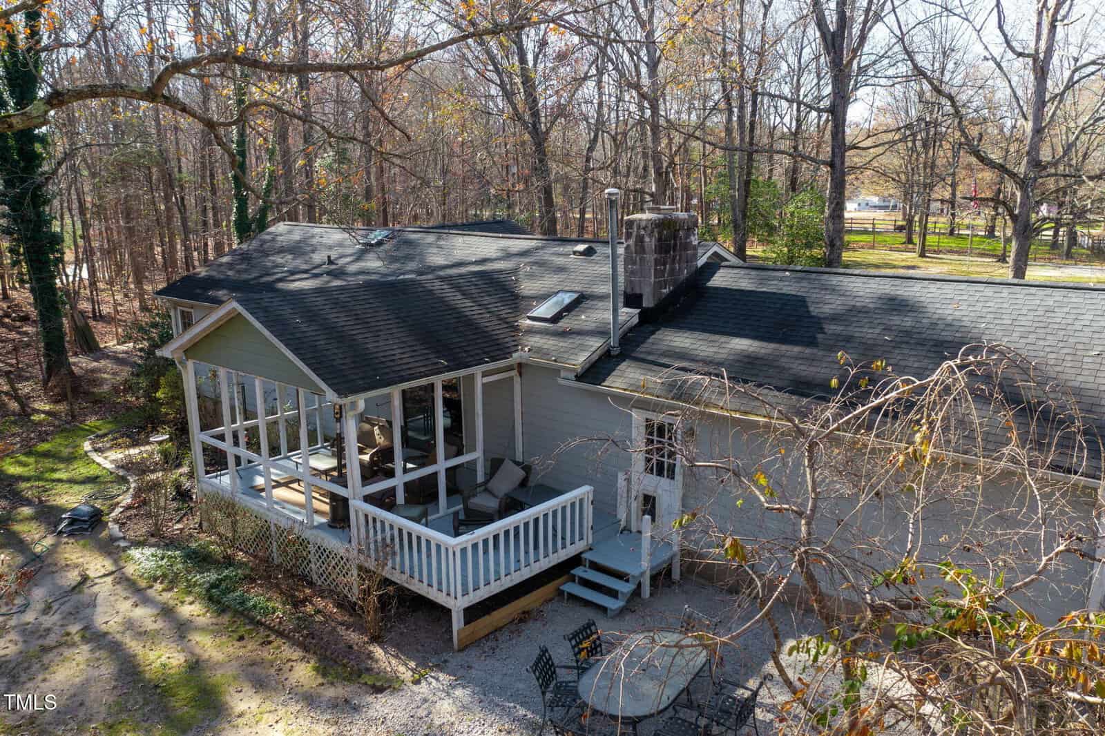 A light gray house with a screened porch, black roof, and small white deck, surrounded by tall trees and a gravel area with a round table and chairs.