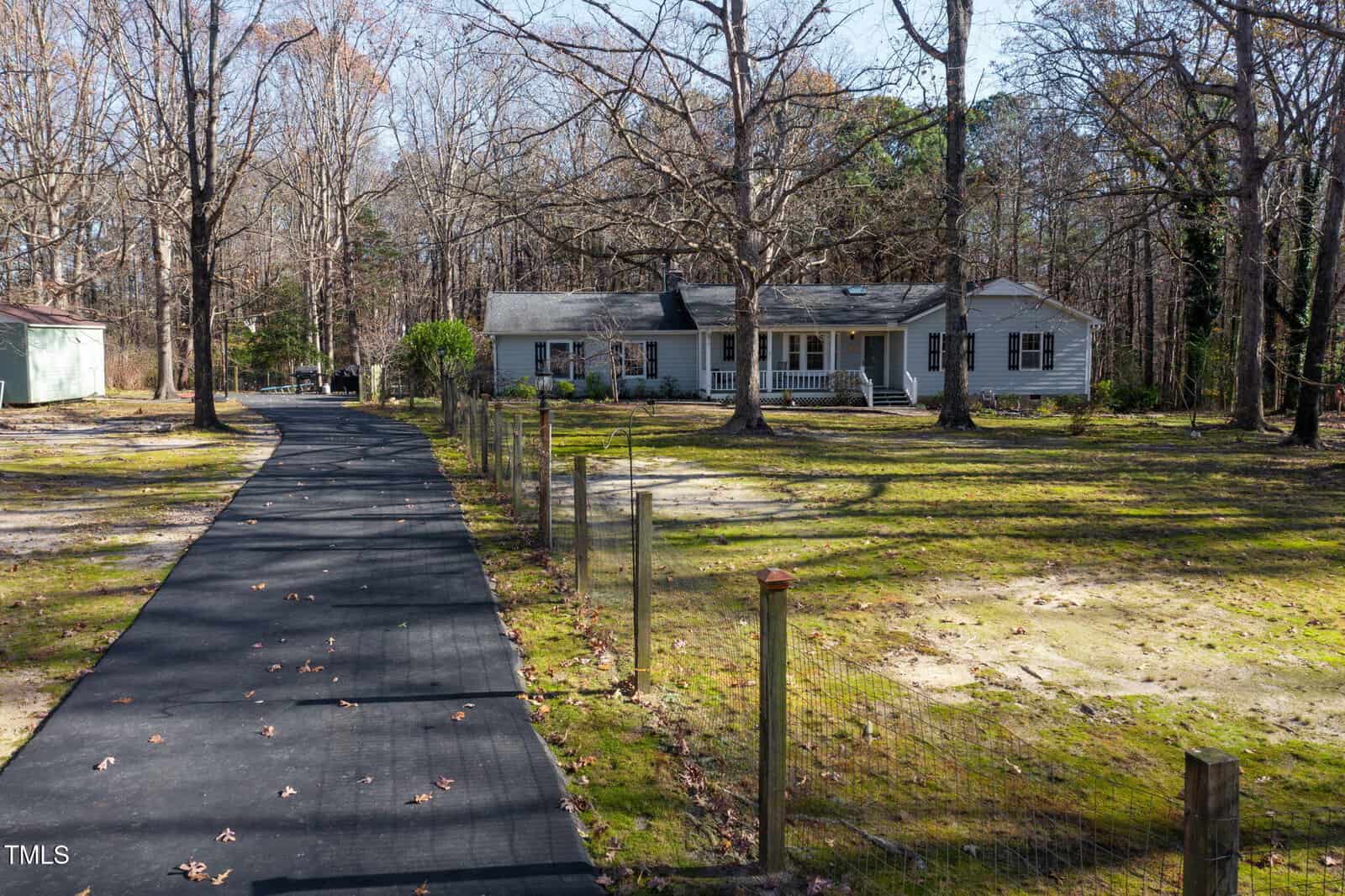 A single-story house with a porch sits among tall trees. A paved driveway leads to the house, and a wire fence lines the yard. The area is partly shaded with patches of grass and bare ground.