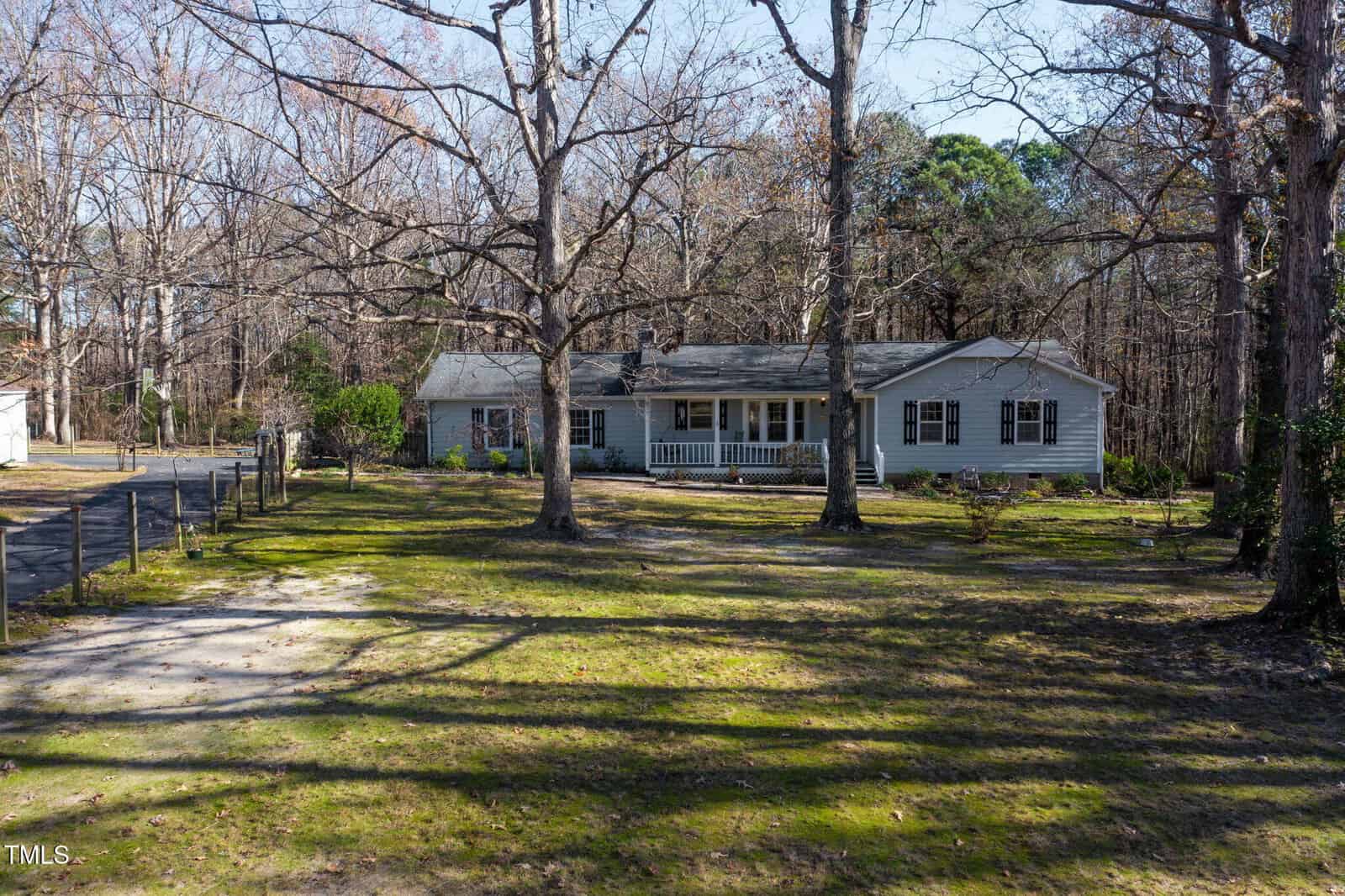 A single-story gray house with white trim sits among tall leafless trees. The yard is spacious with patches of grass and a gravel driveway leading to the house. Shadows from the trees stretch across the lawn.
