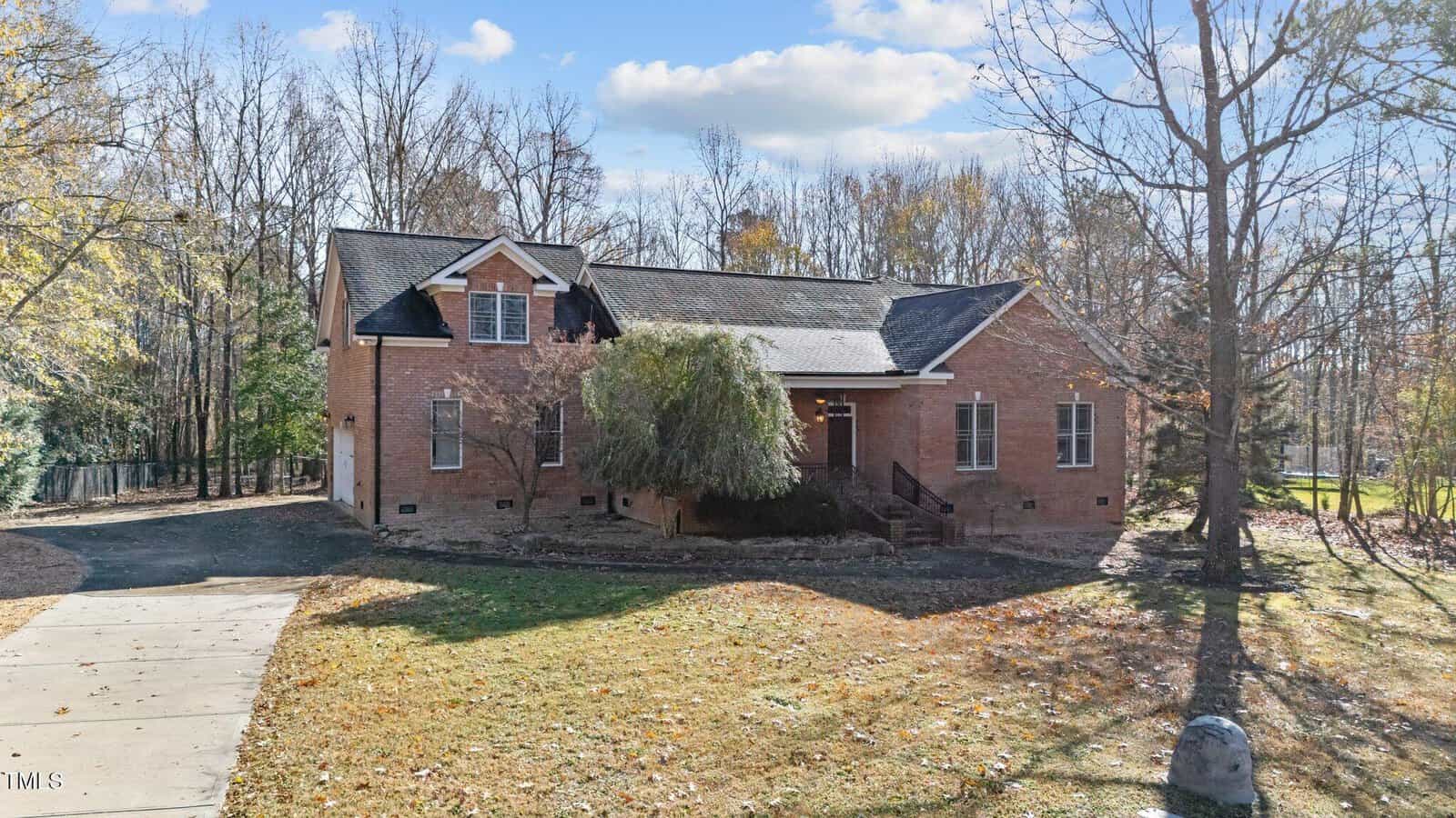 A two-story brick house with a gabled roof and dormer windows, surrounded by leafless trees. A concrete driveway leads to the side of the house, and the front yard is covered with grass and fallen leaves.