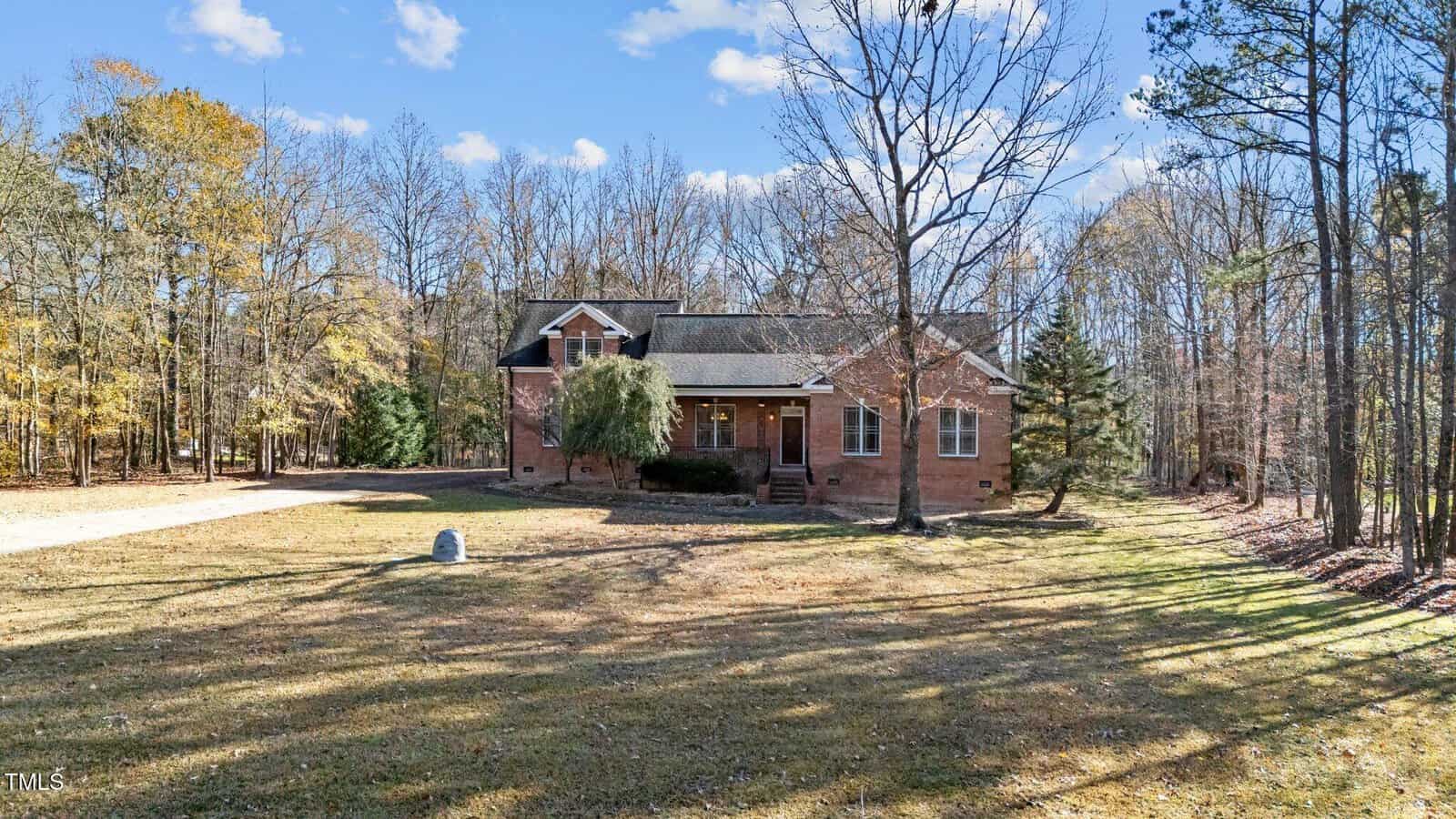 A brick house with a covered front porch sits in a large yard surrounded by trees. The windows are trimmed in white, and a driveway curves to the left side of the home. The sky is mostly clear with a few clouds.