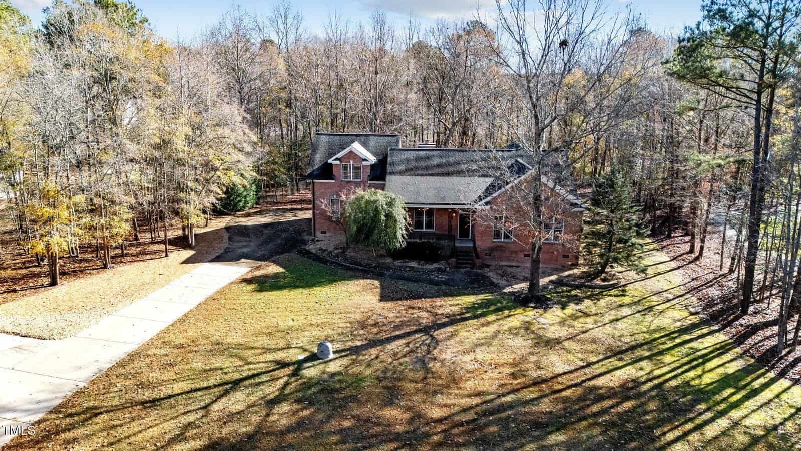 A brick house with a gabled roof sits surrounded by tall trees, with a concrete driveway leading up to it. The front yard is grassy with some leafless trees casting long shadows.
