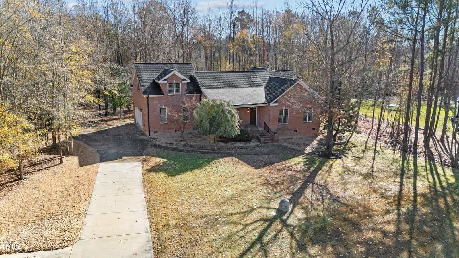 A brick house with a front porch is surrounded by trees and a partially-shaded yard. A concrete driveway leads to the front of the house. The scene appears to be taken in late fall or early winter.
