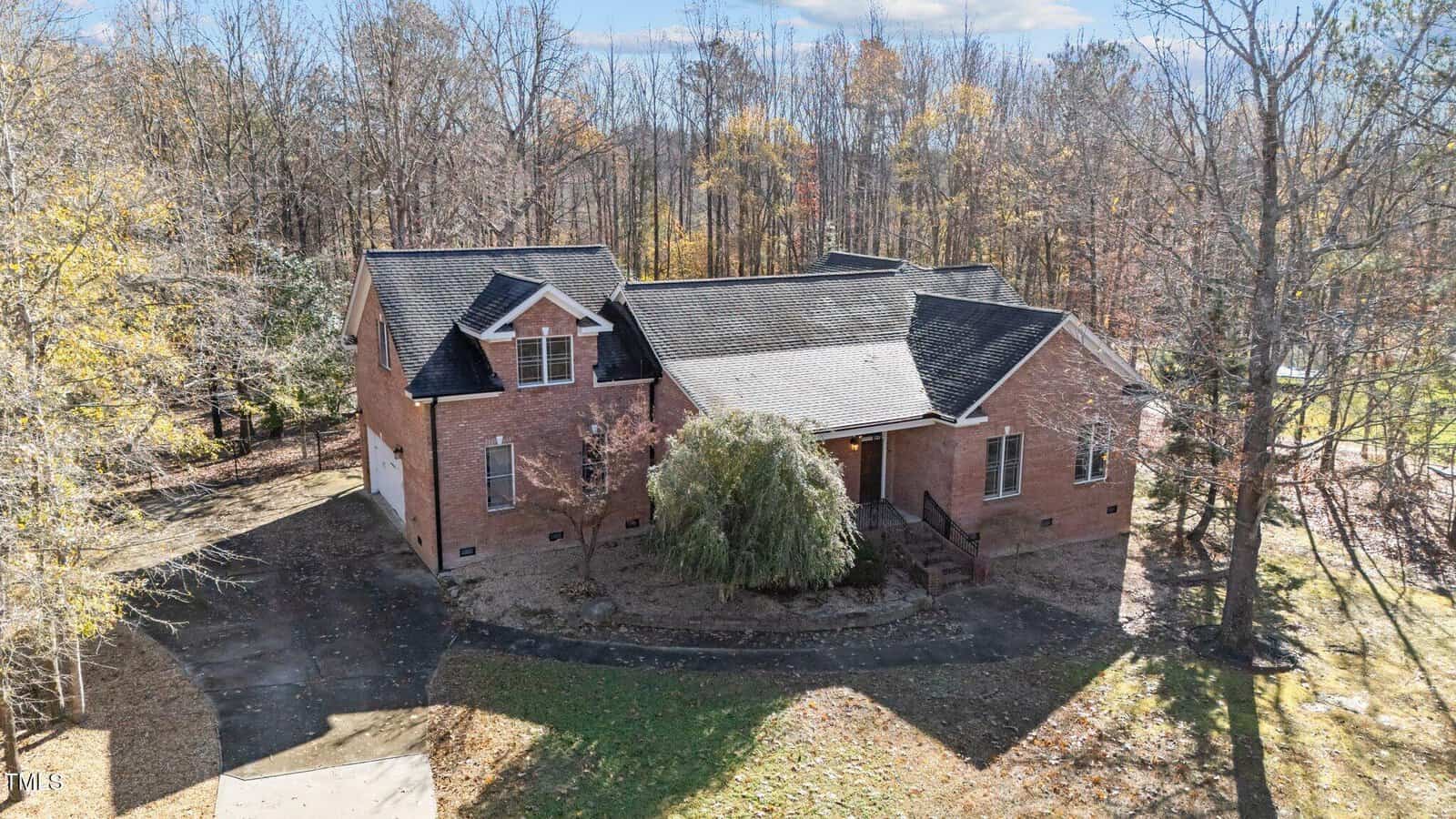 A two-story red brick house with an attached garage, surrounded by trees with autumn foliage. There’s a curved driveway, and sunlight filters through the branches. The yard is lightly covered with fallen leaves.