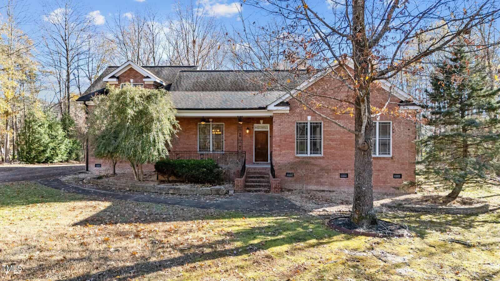 A brick single-story house with a gabled roof, front steps, and a covered entryway, surrounded by trees and a grassy yard on a sunny day.
