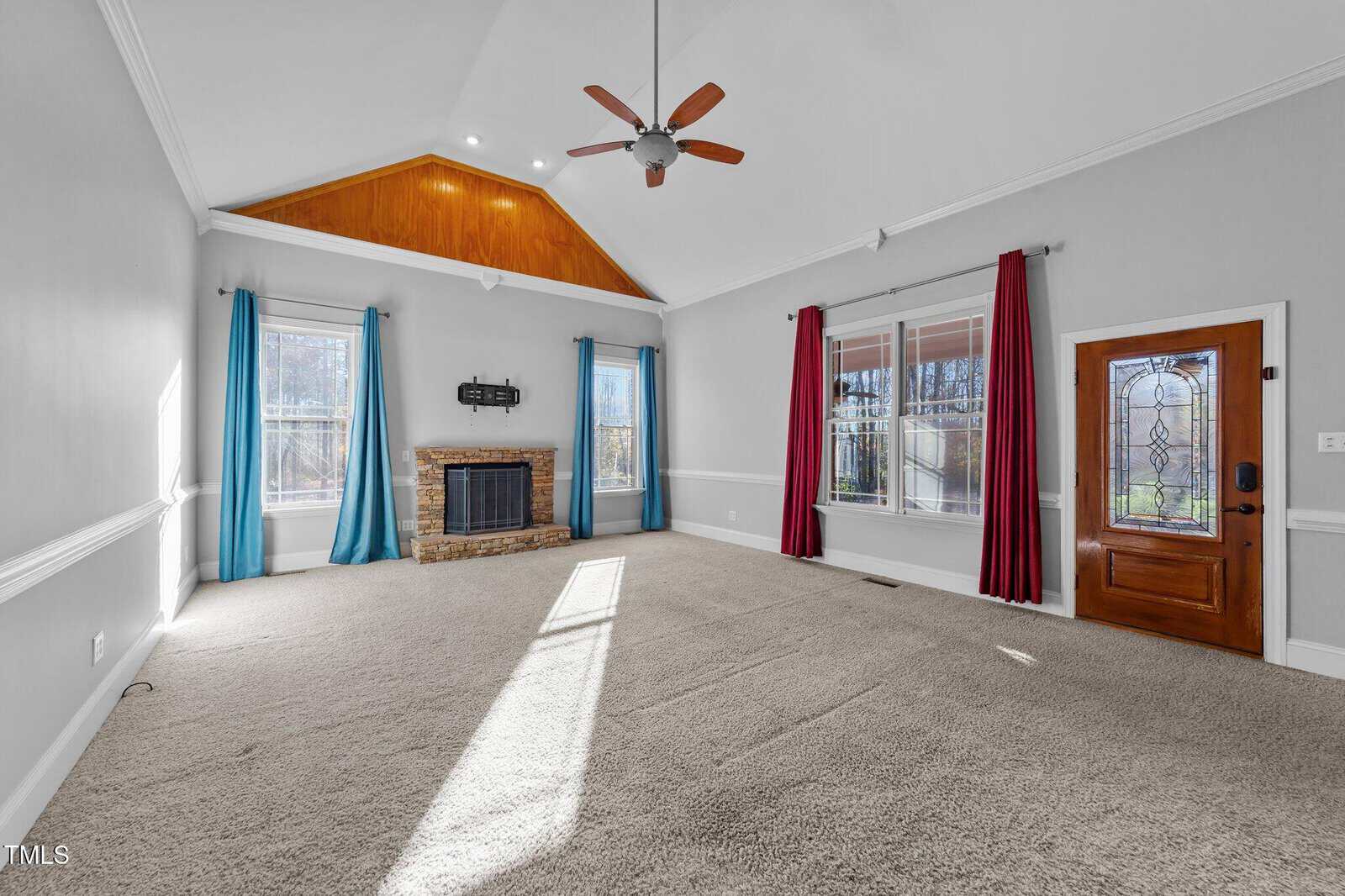 A spacious, carpeted living room with a vaulted ceiling, brick fireplace, two large windows with blue and red curtains, a wooden front door with decorative glass, and a ceiling fan.