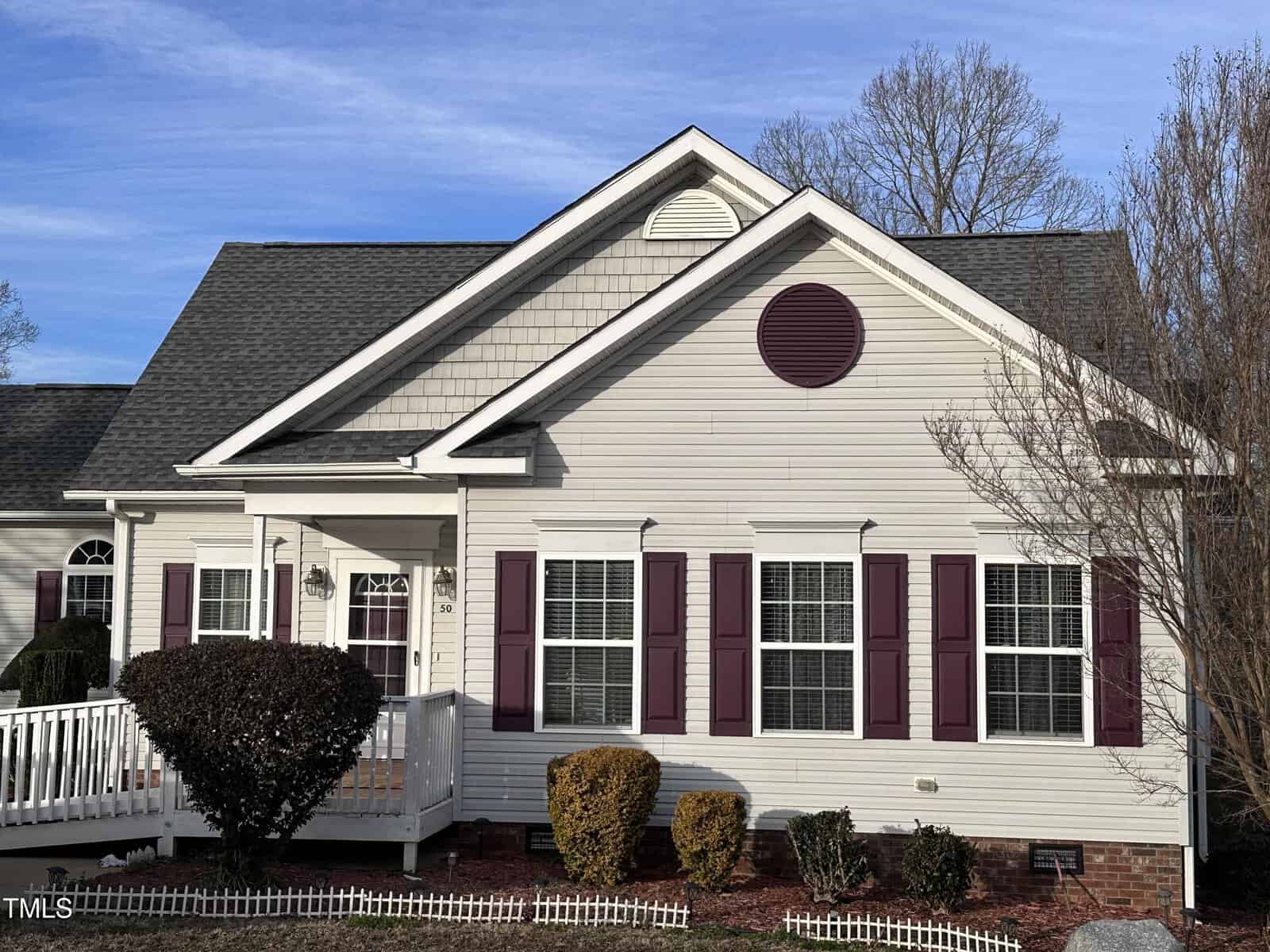 A single-story beige house with maroon shutters and a matching decorative circle above the front windows. There is a small front porch with a white ramp, trimmed bushes, and a fenced yard with mulch and shrubs.