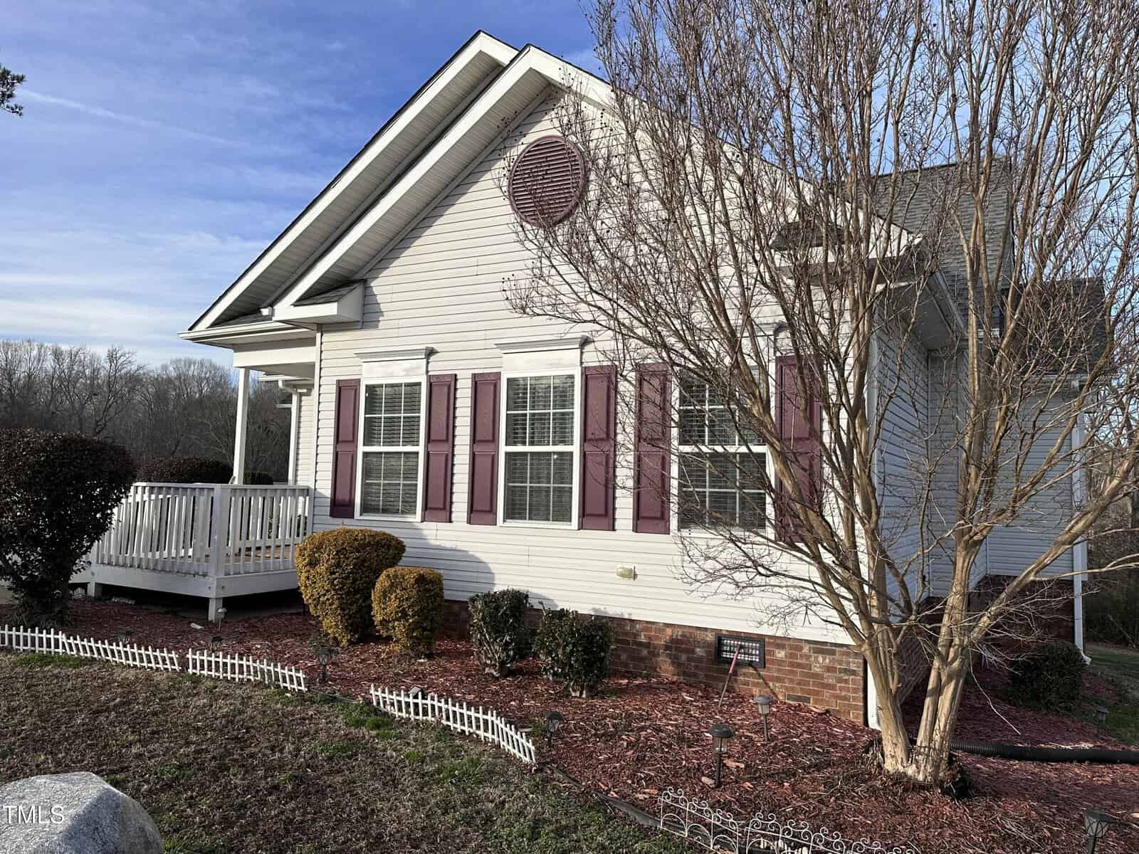 A white house with purple shutters and a small front porch is shown. There is a leafless tree and neatly trimmed bushes in the front yard, with red mulch and a low decorative white fence along the edge.
