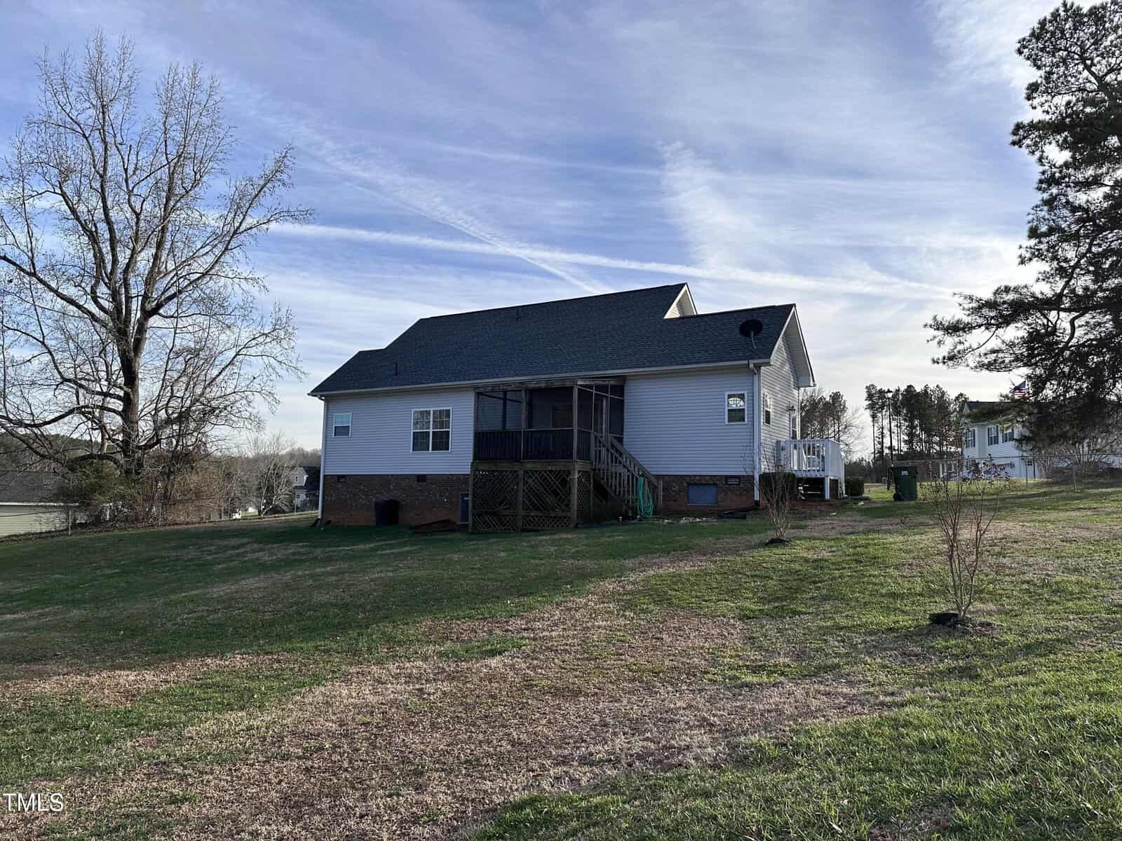 A single-story house with gray siding and a dark roof, featuring a screened porch and a raised wooden deck, sits on a grassy lot with a few trees and open sky in the background.