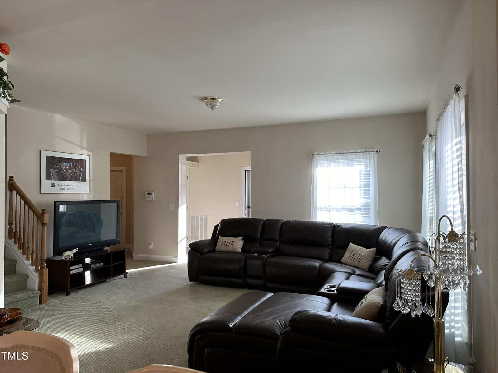 A living room with a large dark sectional sofa, a TV on a stand, a framed picture on the wall, and a staircase with wooden railings. Sunlight enters through windows with white curtains.
