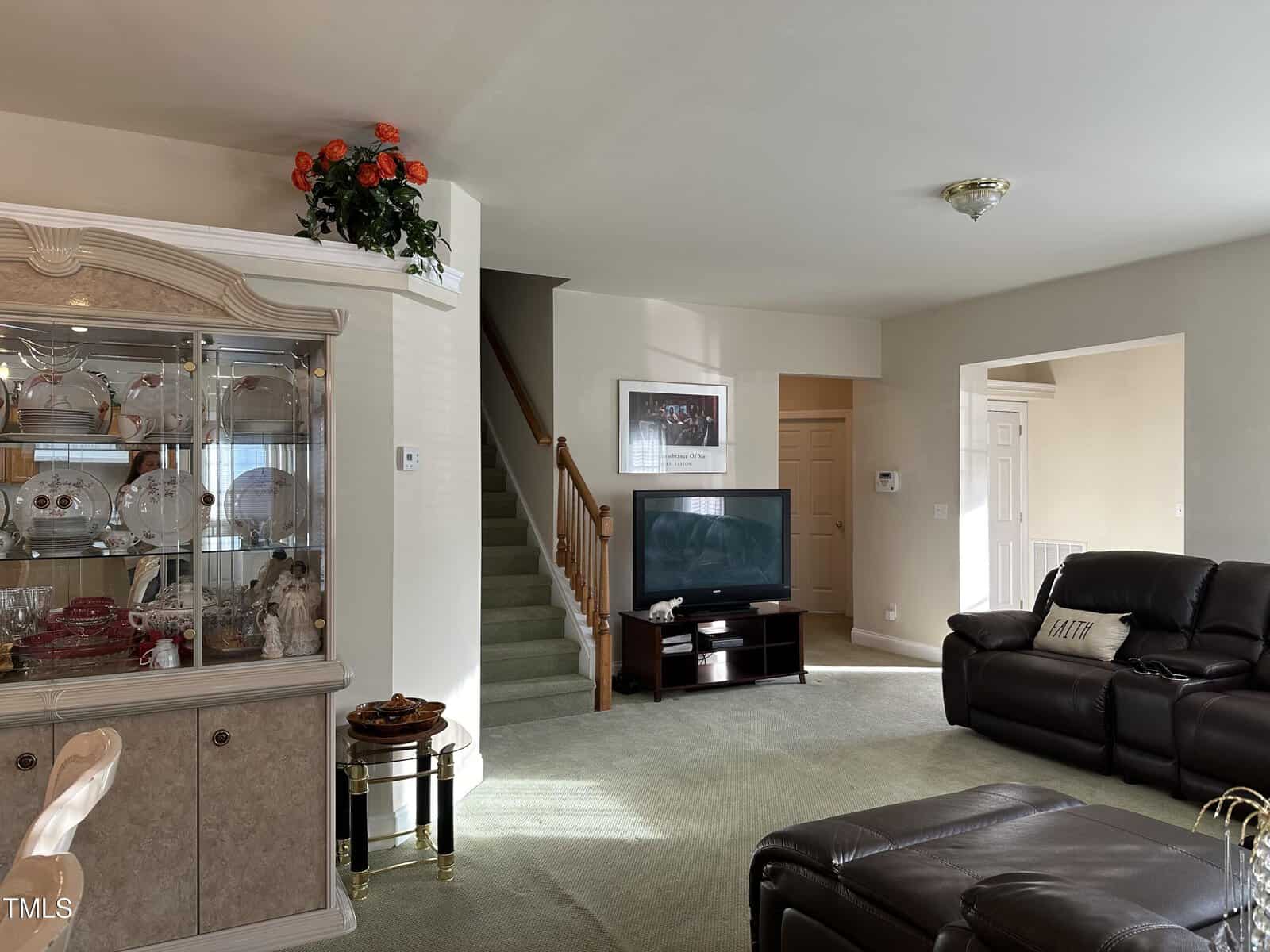 A living room with a green carpet, brown leather sofas, a TV, a china cabinet with dishes, a side table, and stairs leading upward. Sunlight streams in from a window, and red flowers sit atop the cabinet.