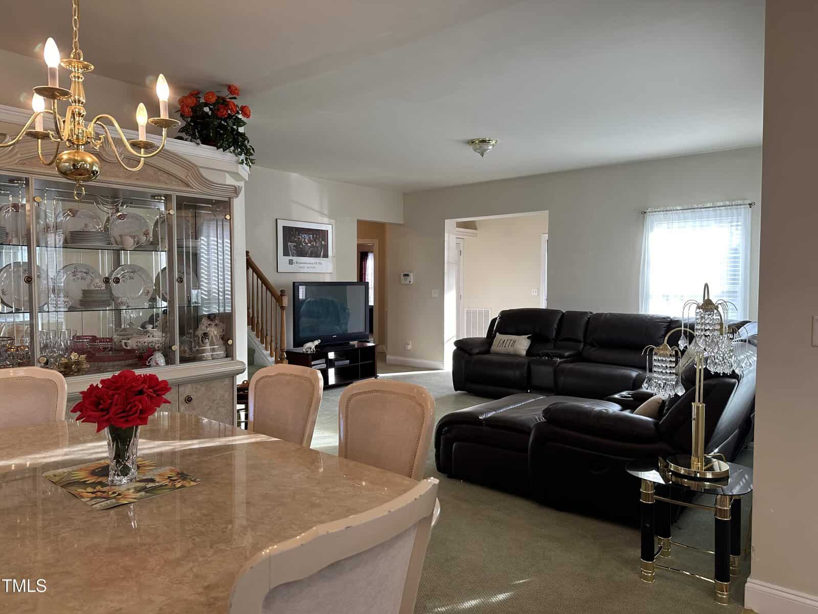A living room with a black sectional sofa, TV, and a glass coffee table. Adjacent dining area features a marble table, cream chairs, and a china cabinet. Bright and cozy ambiance with natural light.