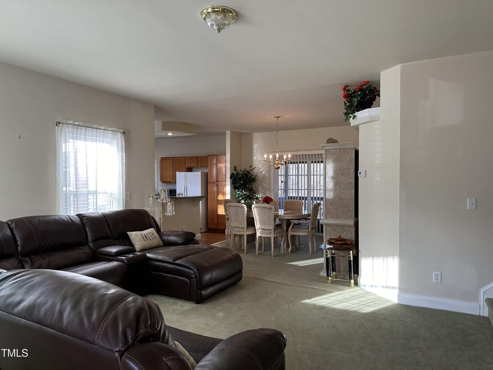 A living room with dark leather sofas, green carpet, and a view into a dining area with a table and six chairs. Beyond the dining room is a kitchen with wooden cabinets and white appliances. Natural light fills the space.