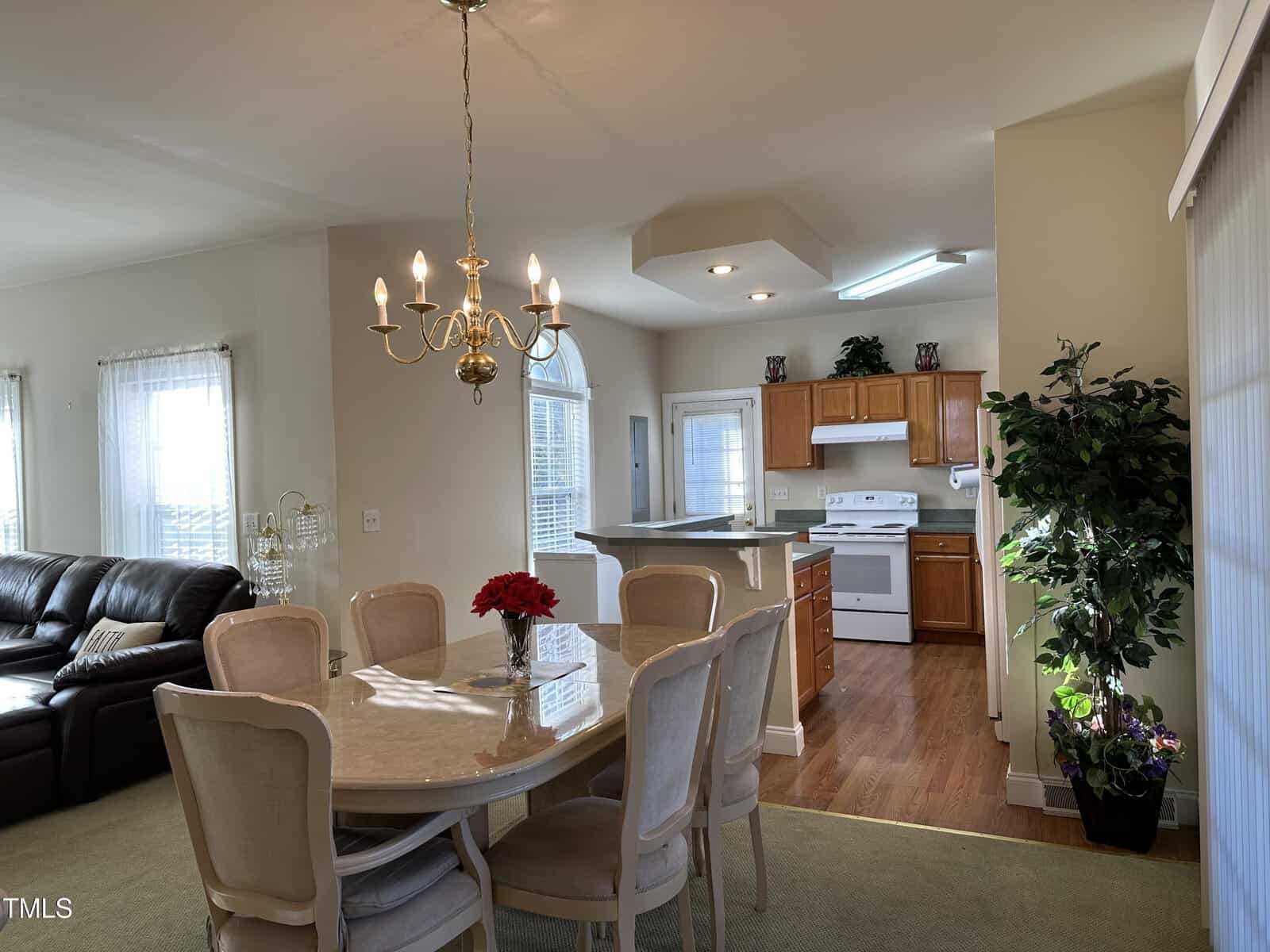 A dining area with a table and six chairs sits beside a bright kitchen with wooden cabinets, white appliances, and a plant in the corner. A chandelier hangs above the table, and sunlight streams through large windows.