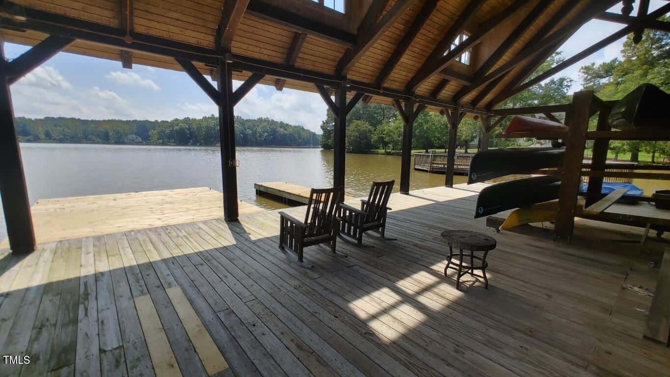 A covered wooden dock with rocking chairs and a fire pit overlooks a calm lake. Canoes are stored on racks to the right, and the area is surrounded by trees under a partly cloudy sky.