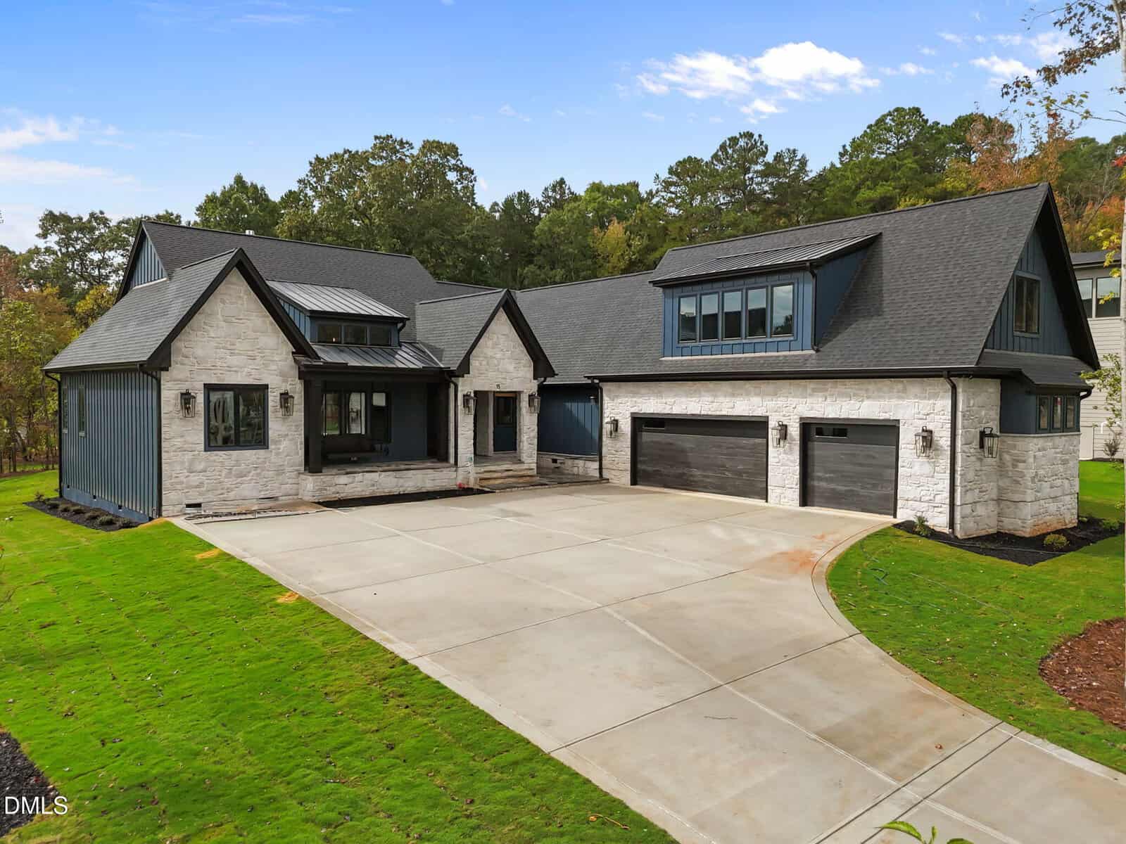 A modern two-story house with light stone exterior, dark trim, and a black shingle roof. The home has a three-car garage and a large driveway, surrounded by a green lawn and trees in the background.