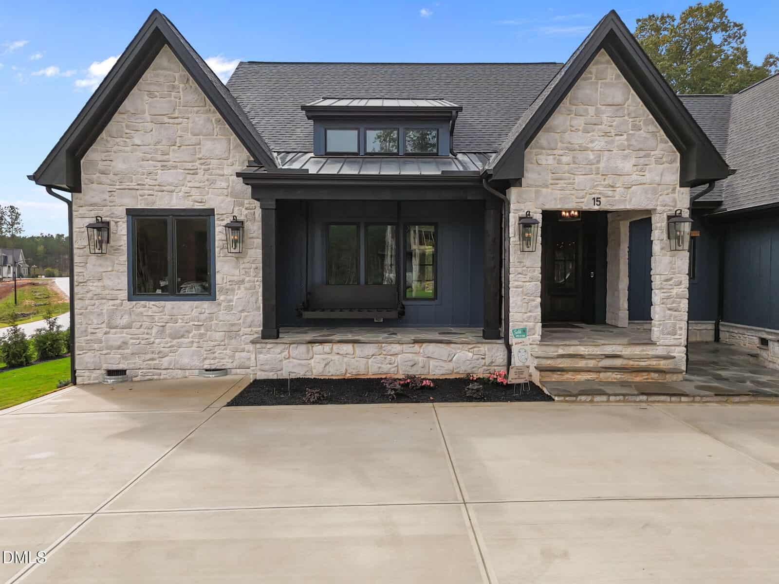 A modern house with light stone exterior, black window frames, a black front door, a porch with two columns, a front step, and a wide concrete driveway leading to the entrance.