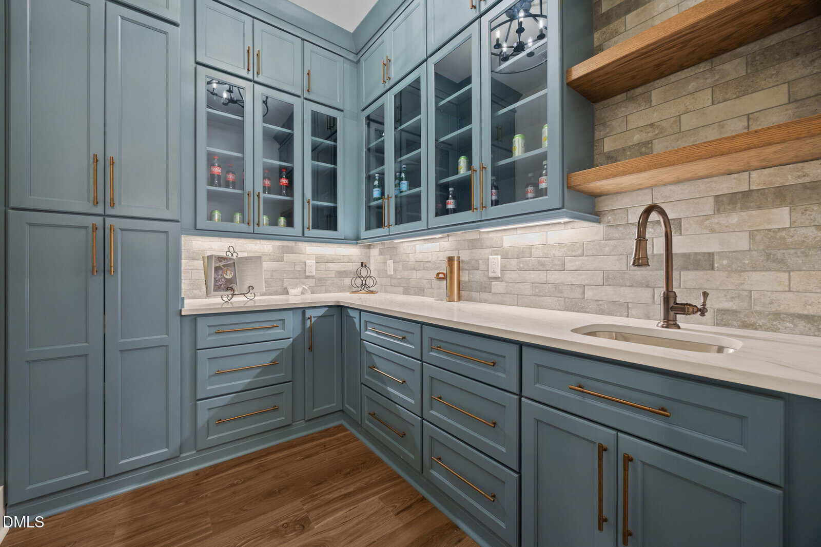 Modern kitchen with blue cabinets, gold hardware, white countertops, glass cabinet doors, wood shelves, tile backsplash, and wood flooring. Recessed lighting illuminates the space.
