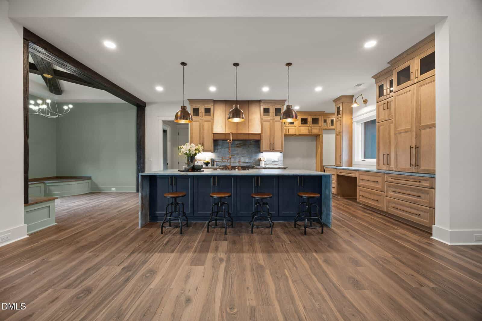 Spacious modern kitchen with wood floors, light wood cabinets, a large blue island with four stools, pendant lights, and stainless steel appliances. An open doorway leads to a green room with a chandelier.