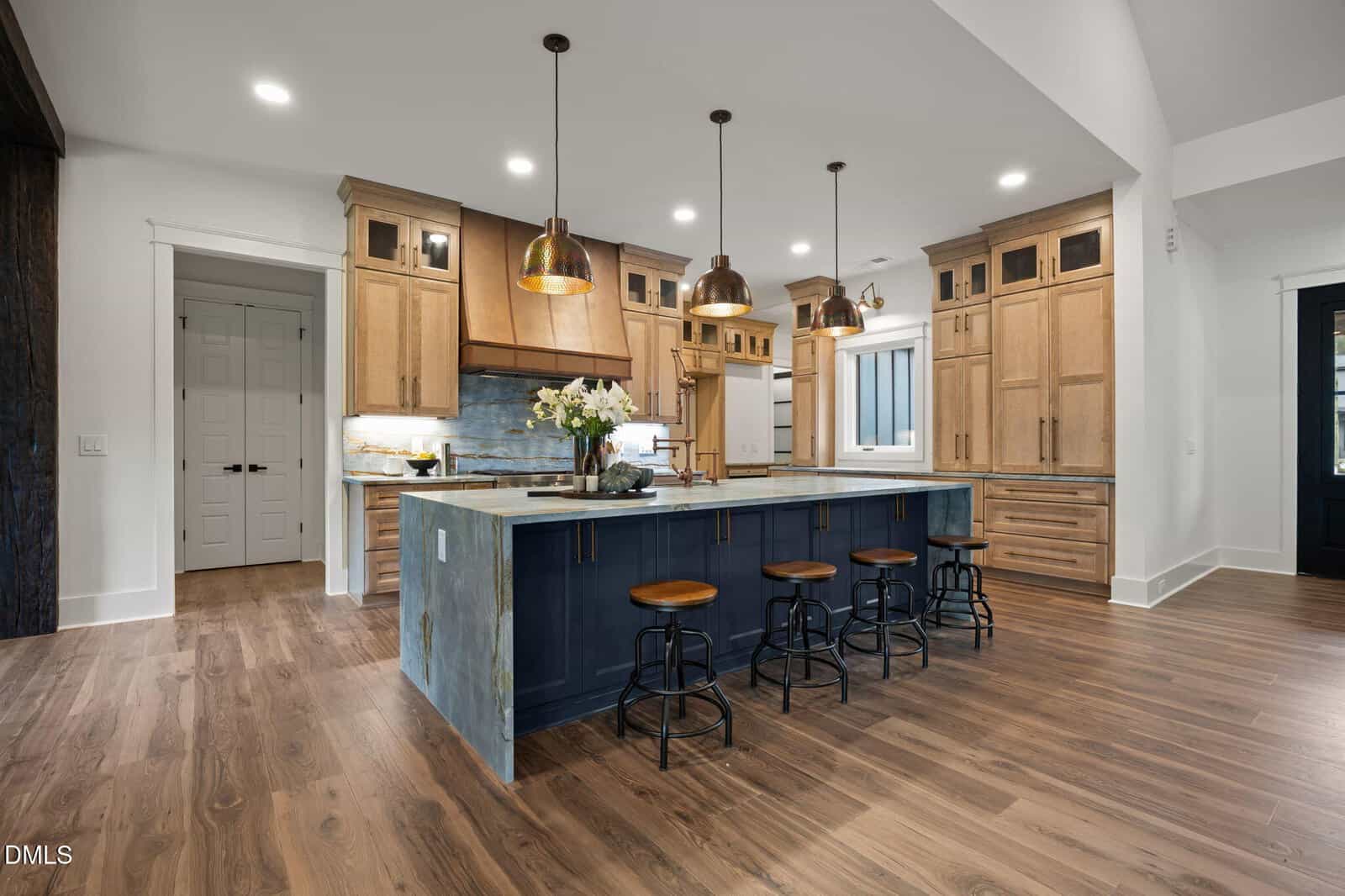 A modern kitchen with wood cabinets, a blue island with a stone countertop, four metal stools, pendant lights, light wood flooring, and stainless steel appliances. A vase with flowers sits on the island.