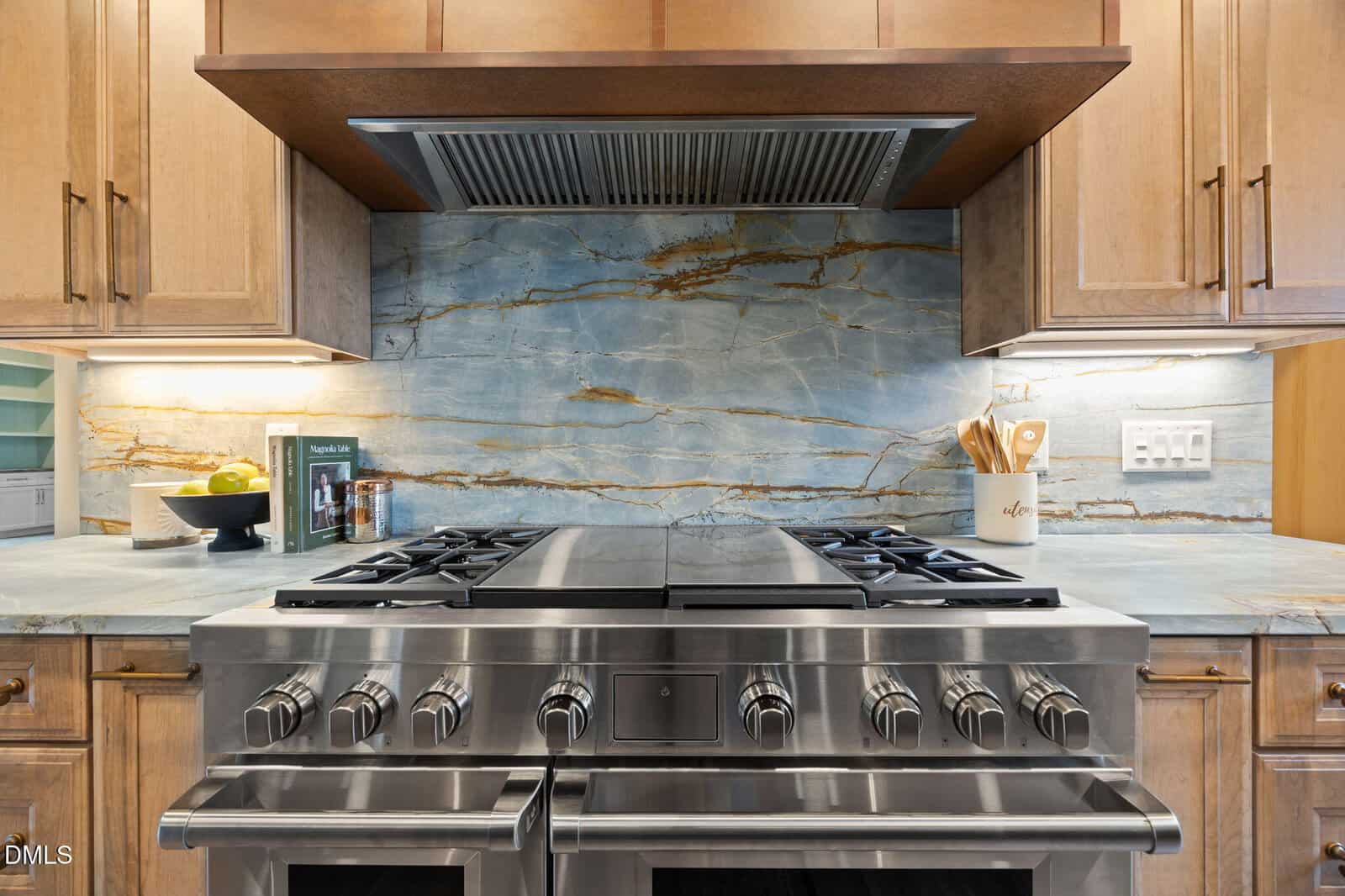 A modern stainless steel gas stove with two ovens, set against a blue and beige marble backsplash. Wooden cabinets surround the stove, and kitchen utensils and a bowl of fruit are on the countertop.