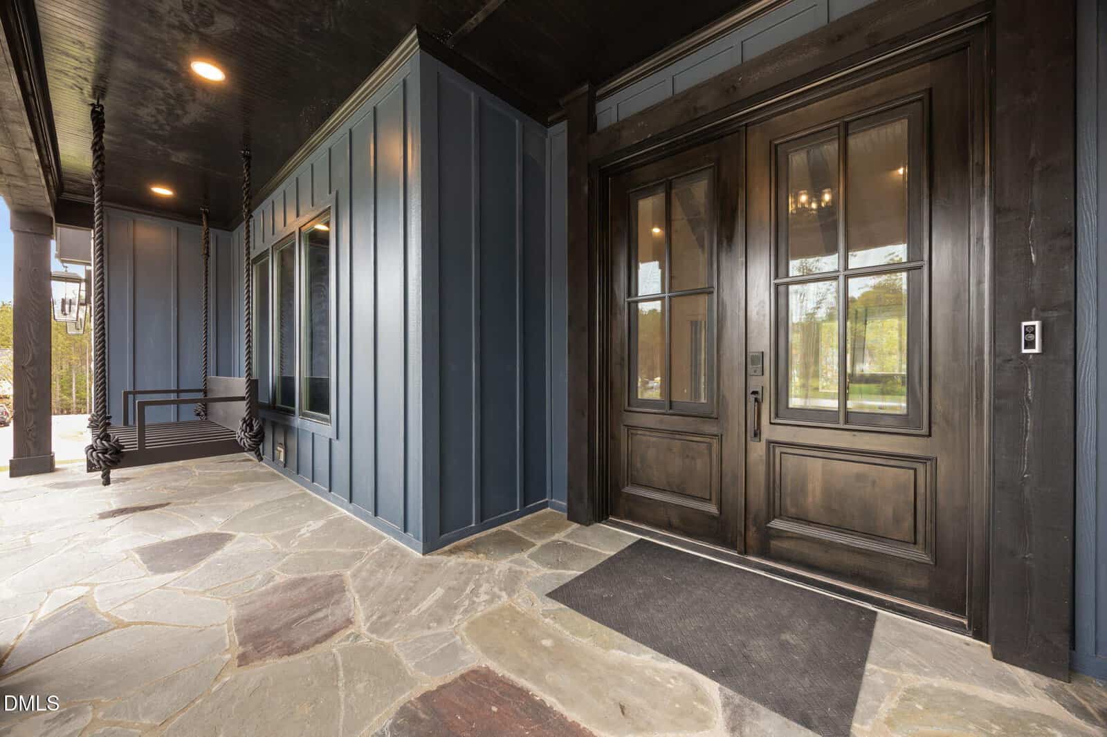 A front porch with a stone floor, dark double doors with glass panes, a black porch swing, vertical siding, and a black ceiling with recessed lights. A doormat is placed in front of the doors.