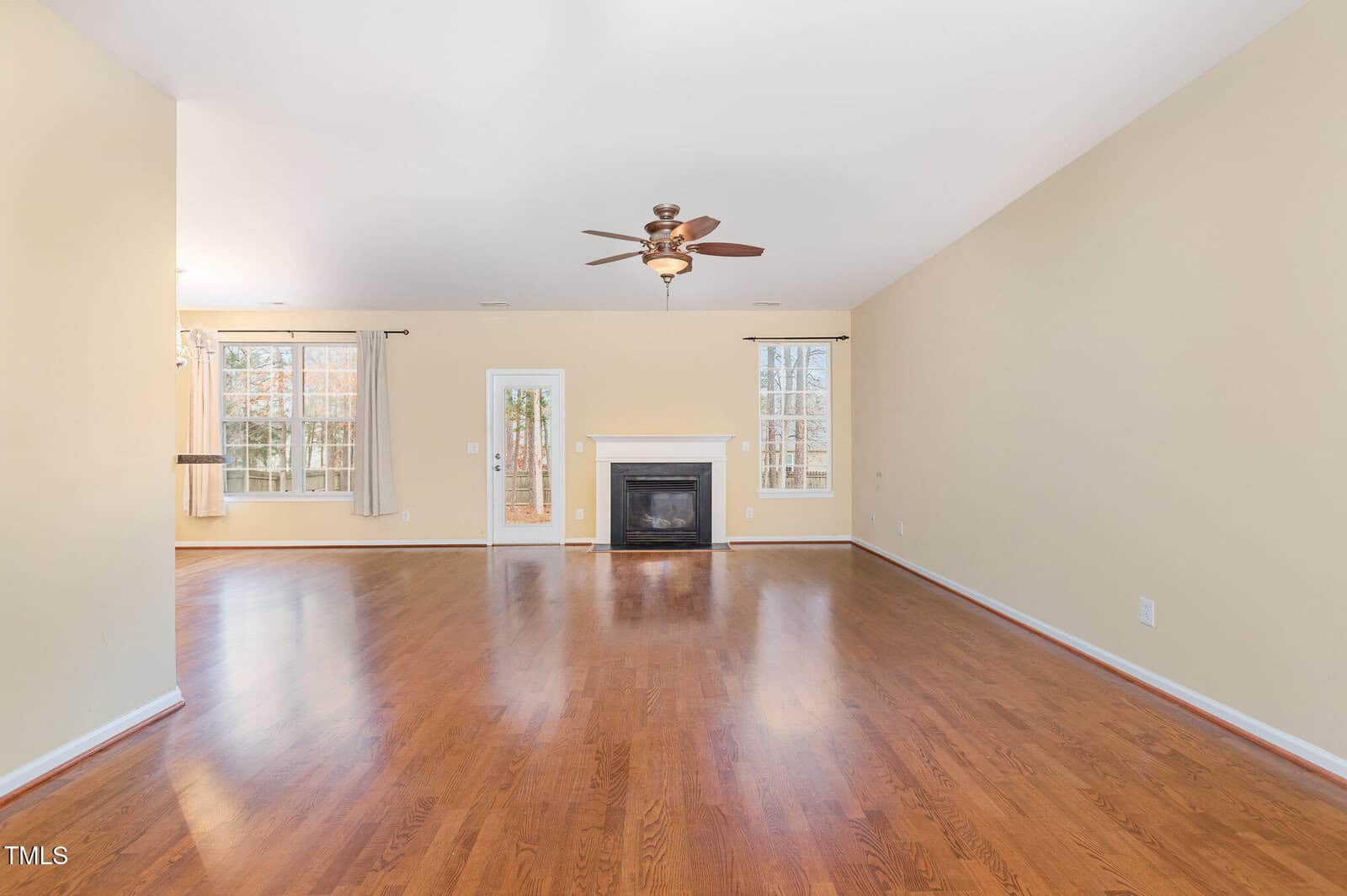 A spacious, empty living room with light-colored walls and hardwood floors. It features a ceiling fan with a light fixture, a central fireplace, and large windows. French doors lead to the outside, allowing natural light to enter the room.