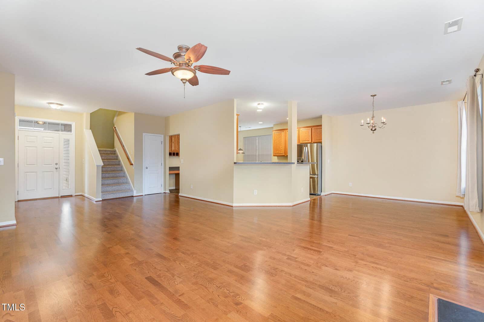 Spacious living area with hardwood flooring, a ceiling fan, and natural light. The open layout features a kitchen with a breakfast bar and stainless steel appliances, a dining area with a chandelier, and stairs leading to the second floor.