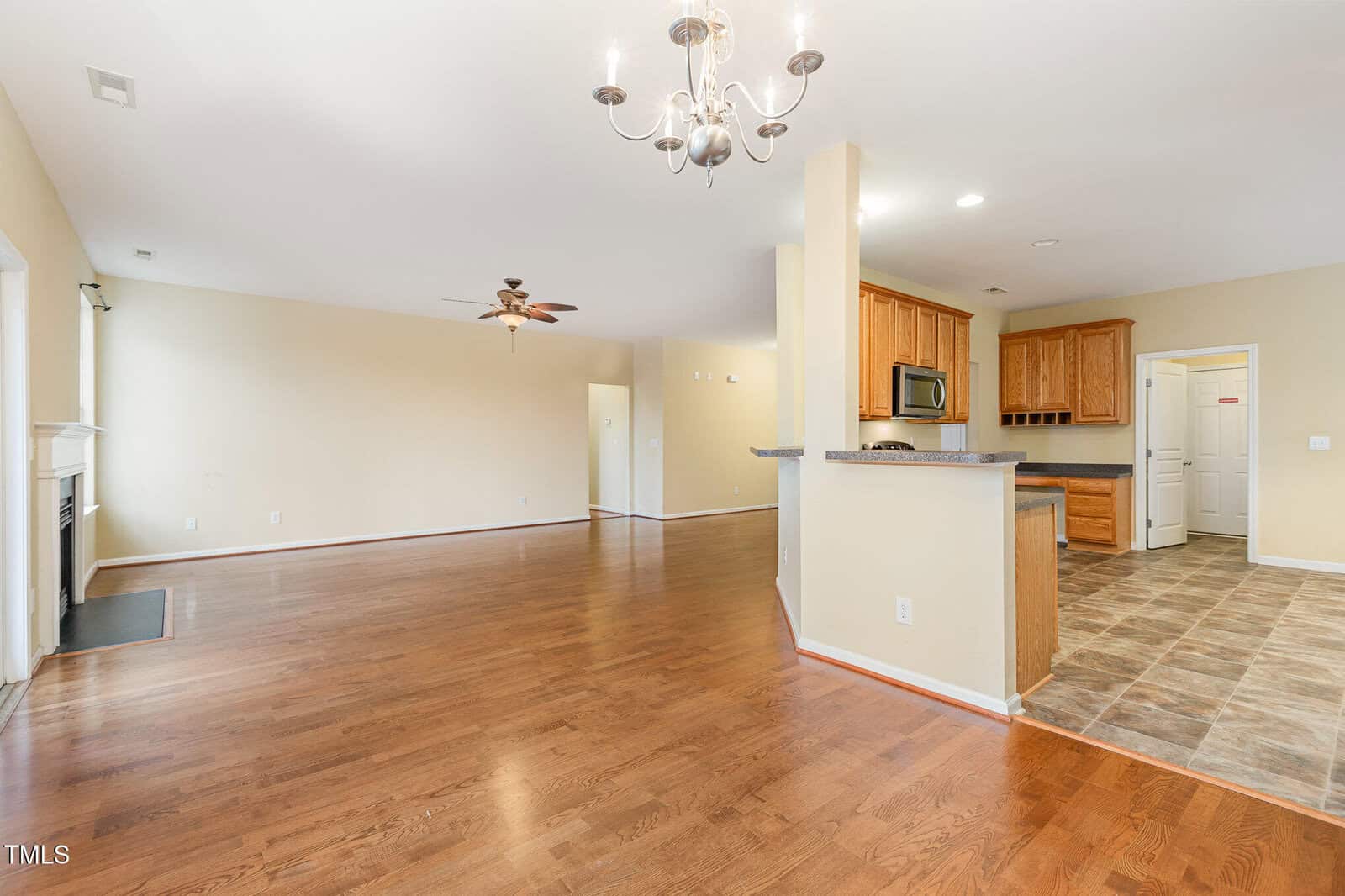 A spacious open-concept living room and kitchen with hardwood floors. The kitchen has wooden cabinets, a stainless steel microwave and oven, and a gray countertop. A chandelier hangs from the ceiling and a ceiling fan is in the living area.