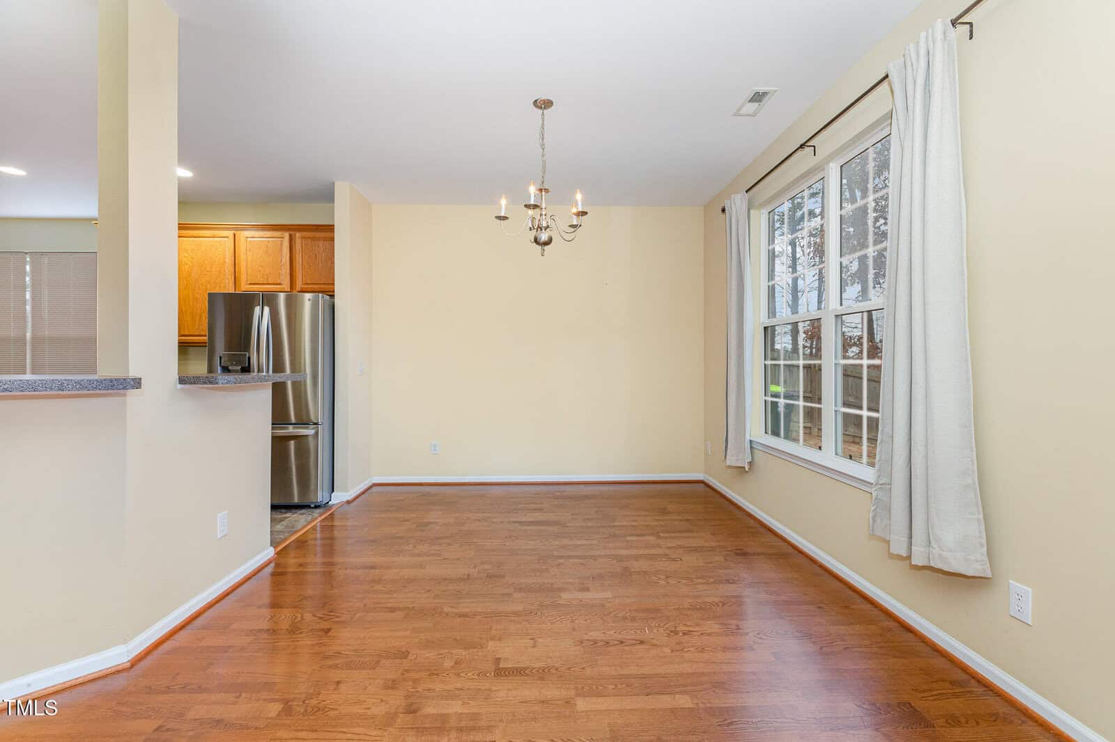 An empty room with wooden floors and beige walls. A chandelier hangs from the ceiling. Large windows on the right with white curtains let in natural light. A kitchen area with stainless steel appliances is visible on the left.