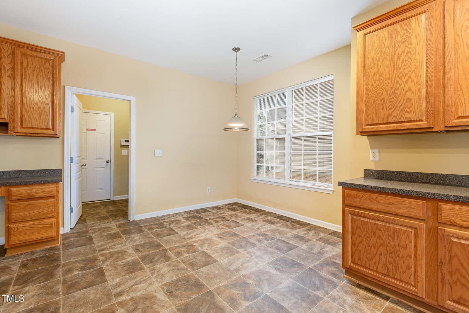 Kitchen with wooden cabinets, dark countertops, and tiled floor. A large window on the right lets in natural light. A hanging light fixture is above a dining area, and a door leads to another room. Walls are painted in a warm beige color.