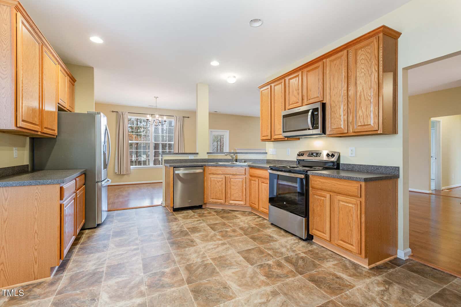 A spacious kitchen features wooden cabinets, dark countertops, stainless steel appliances including a stove and dishwasher, and tile flooring. A windowed area with curtains and a glimpse of an adjoining room can be seen in the background.