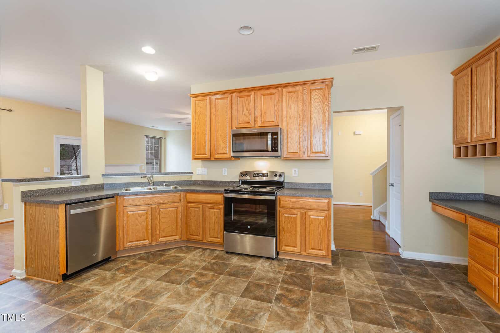 A spacious kitchen with light wooden cabinets, a stainless steel oven, microwave, and dishwasher. The room features a tile floor and neutral walls. There is a countertop with a sink, open shelving, and an entrance leading to another area.
