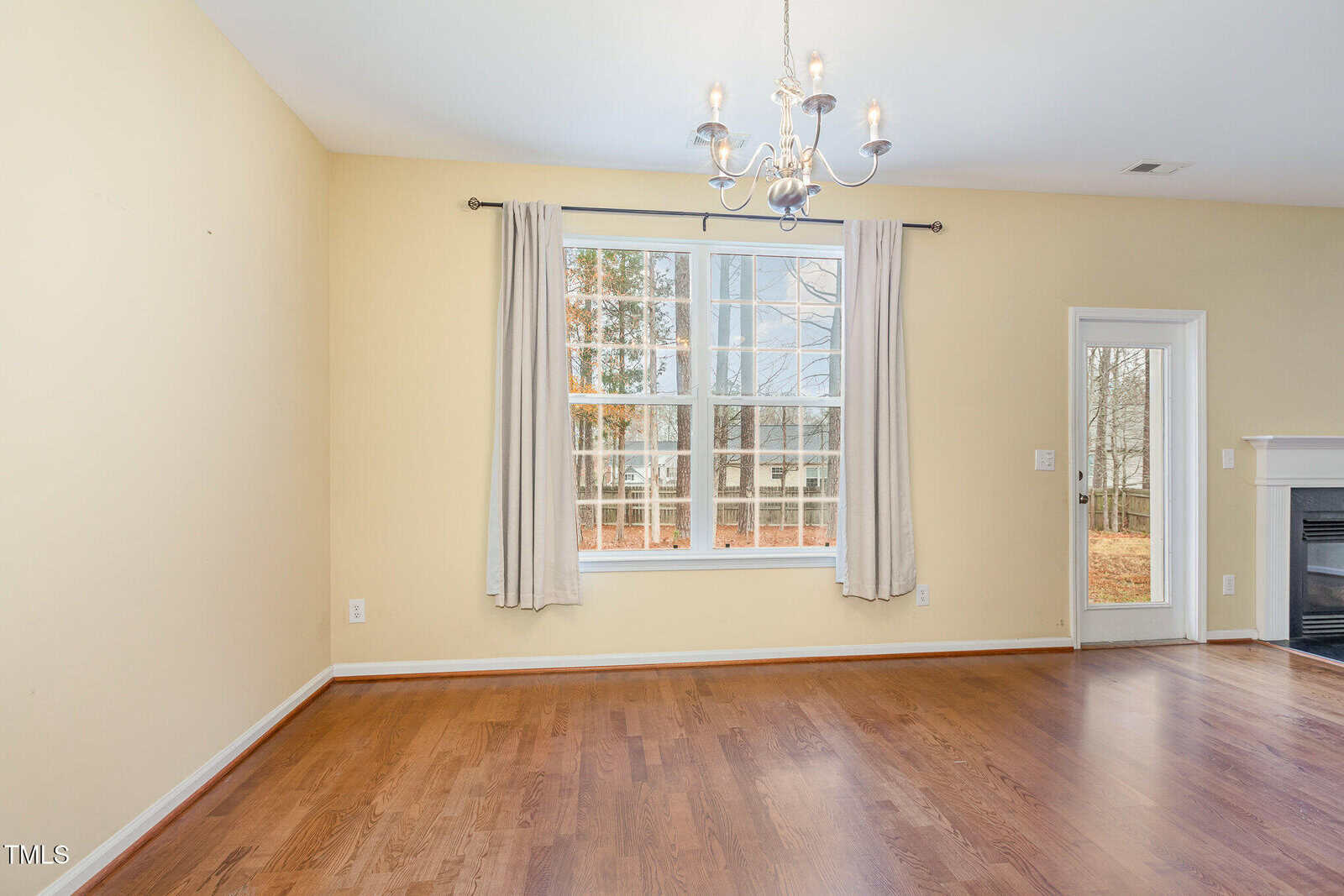 An empty, well-lit room with light yellow walls and wooden floors. A large window with beige curtains offers an outdoor view. A white door is to the right, and a chandelier hangs from the ceiling. A fireplace is partially visible on the far right.