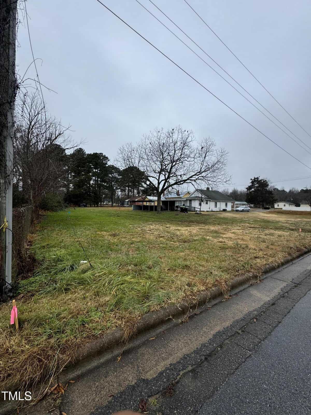 The image captures an overcast day at 0 Woodson Street, where a grassy lot and a few trees stand quietly. A white house with a porch rests in the background, typical of Oxford charm. A paved road runs alongside, and overhead utility lines cut through the gray sky, adding to the scene's simplicity.