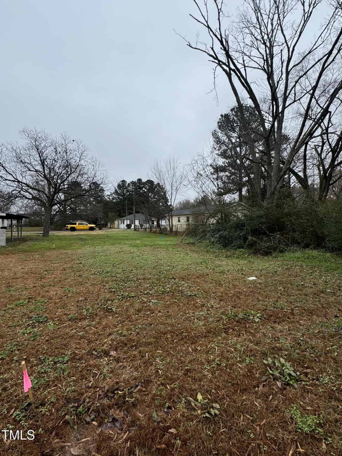 A residential area with a grassy lot and some trees. There is a house in the background, partially obscured by trees. A yellow car is parked near the house. The sky is overcast, and a pink marker is visible in the foreground.
