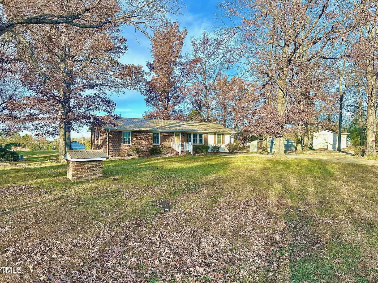 A brick house on 1049 Jackson Royster Road, surrounded by tall trees with autumn foliage. The front yard is covered with fallen leaves, and a small brick structure is near the center. A driveway leads to a garage on the right side under Henderson's clear blue sky.
