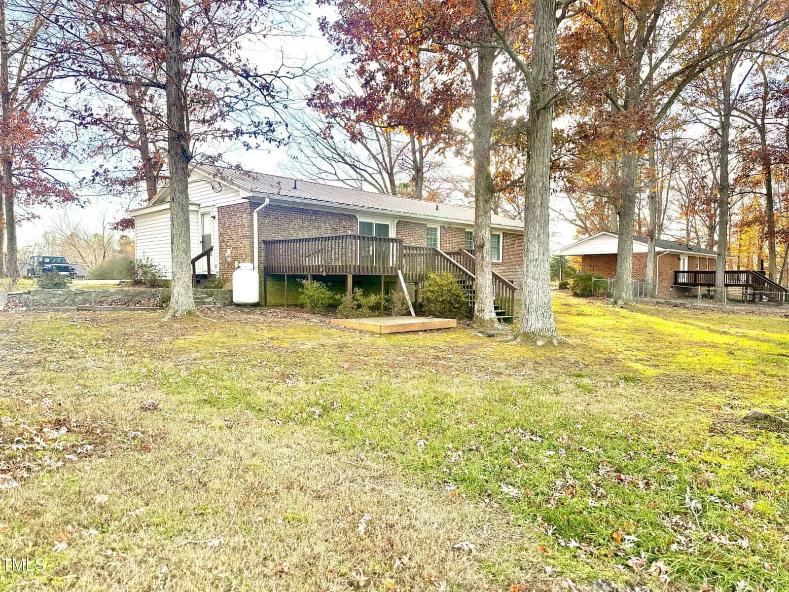A brick house with a wooden deck surrounded by trees with autumn leaves. The grassy yard is sunlit, and another house is visible in the background.