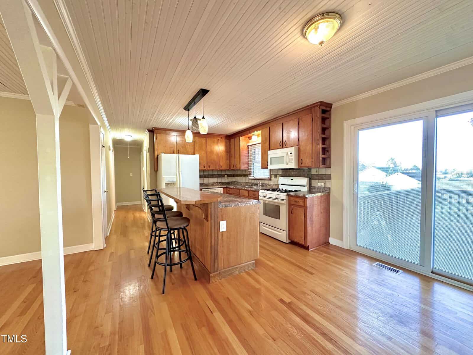 A cozy kitchen featuring wooden cabinets, a white refrigerator, white stove, and a wooden island with bar stools. Sunlight streams through a sliding glass door, highlighting the light hardwood floor and beamed ceiling.